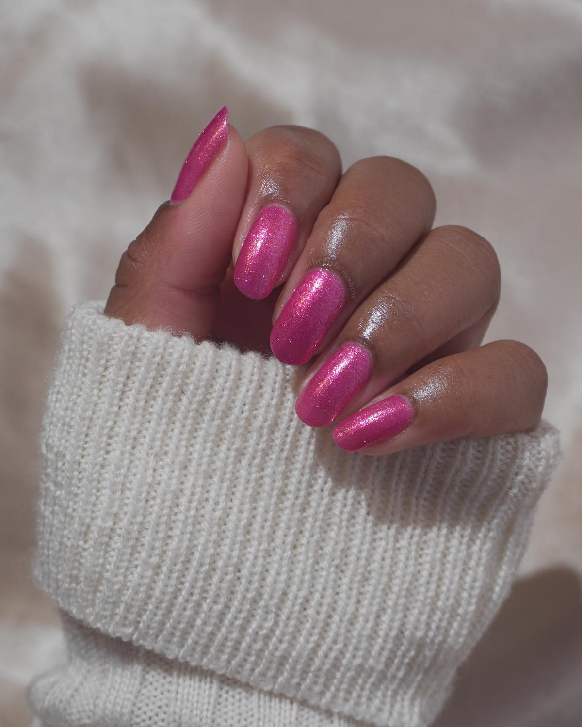 A close-up of a hand with manicured nails painted in a sparkling metallic pink polish, wearing a white ribbed knit sweater cuff against a soft beige background.