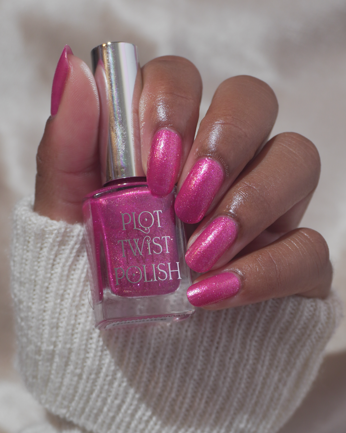 A hand with bright pink glitter nail polish holding a bottle of Plot Twist Polish in the same shade, set against a soft white knit sleeve and light background.