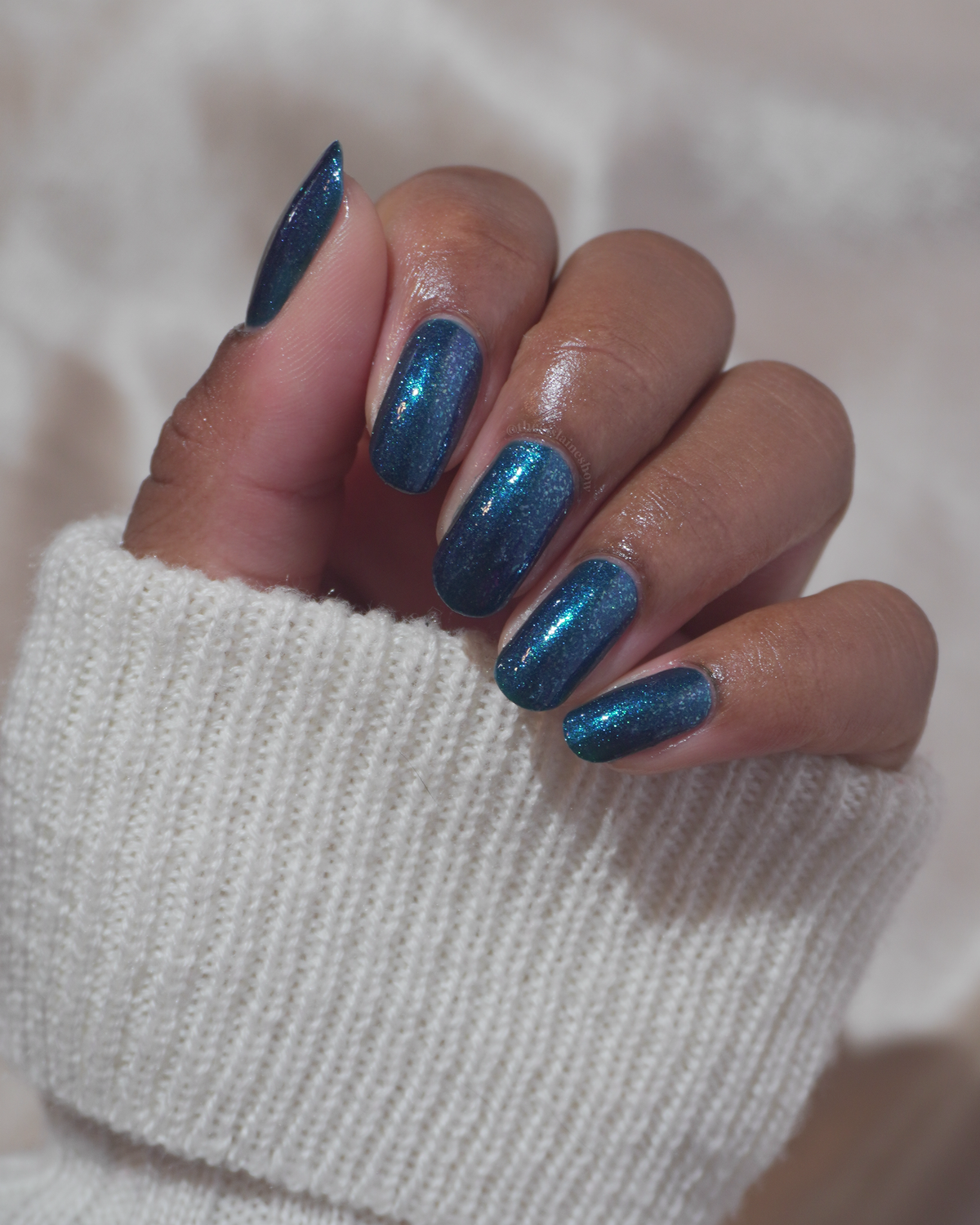 A close-up of a hand with manicured nails painted in a shimmering metallic teal-blue polish, wearing a white knit sweater against a soft neutral background.