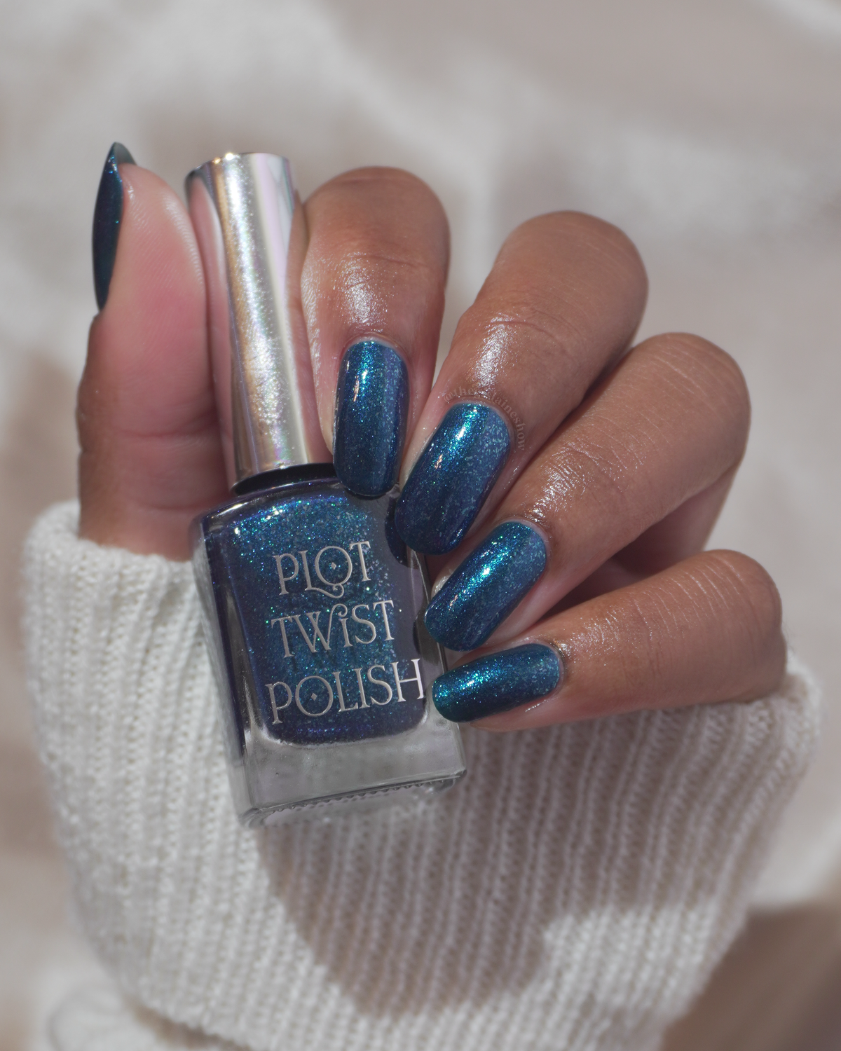 A hand holding a bottle of Plot Twist Polish nail polish, showing the blue glitter polish both in the bottle and applied to the fingernails, against a soft white knit sleeve and light background.