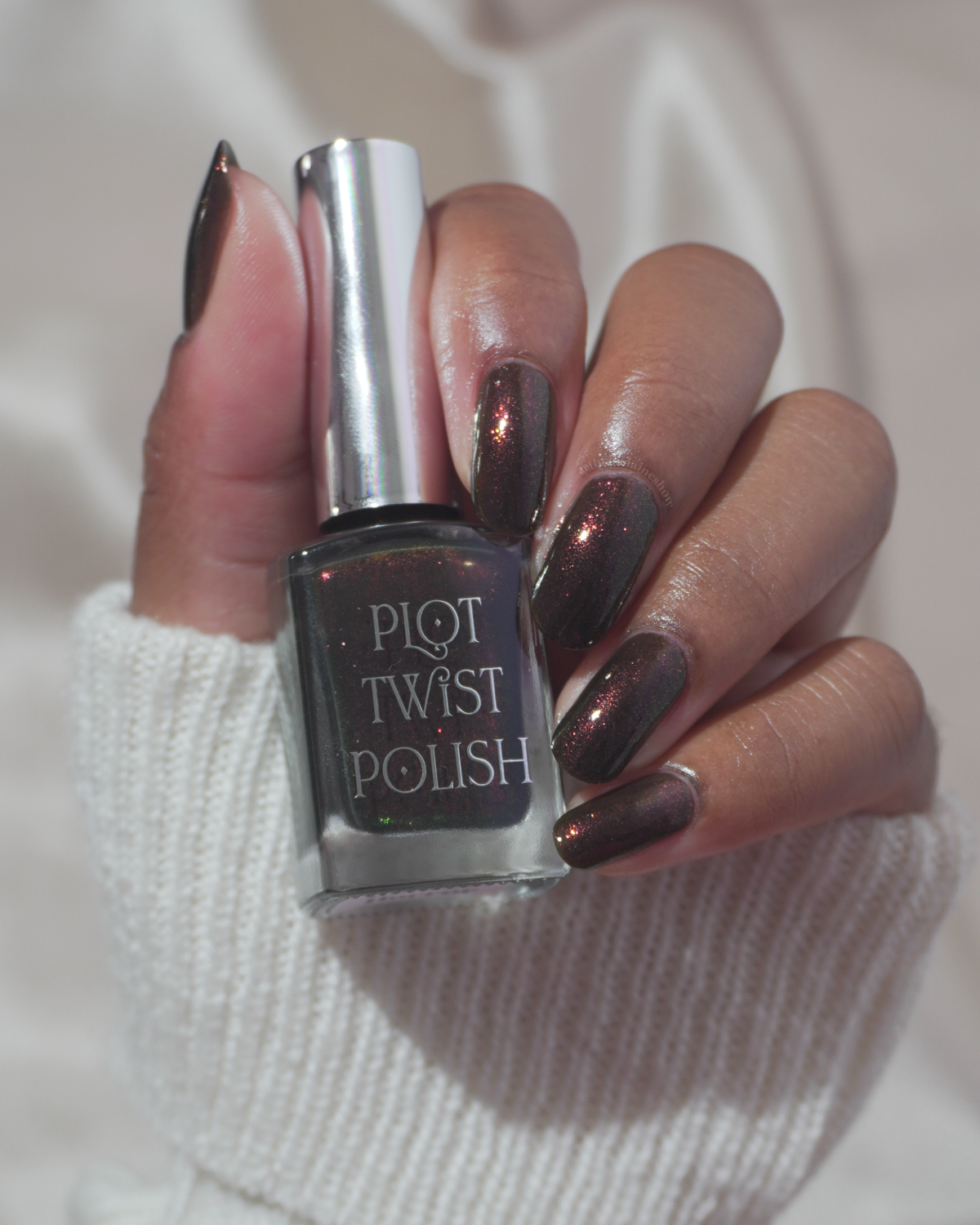 A hand holding a bottle of Plot Twist Polish nail polish, showing the dark metallic bronze shade applied to the nails against a soft white knit sleeve.
