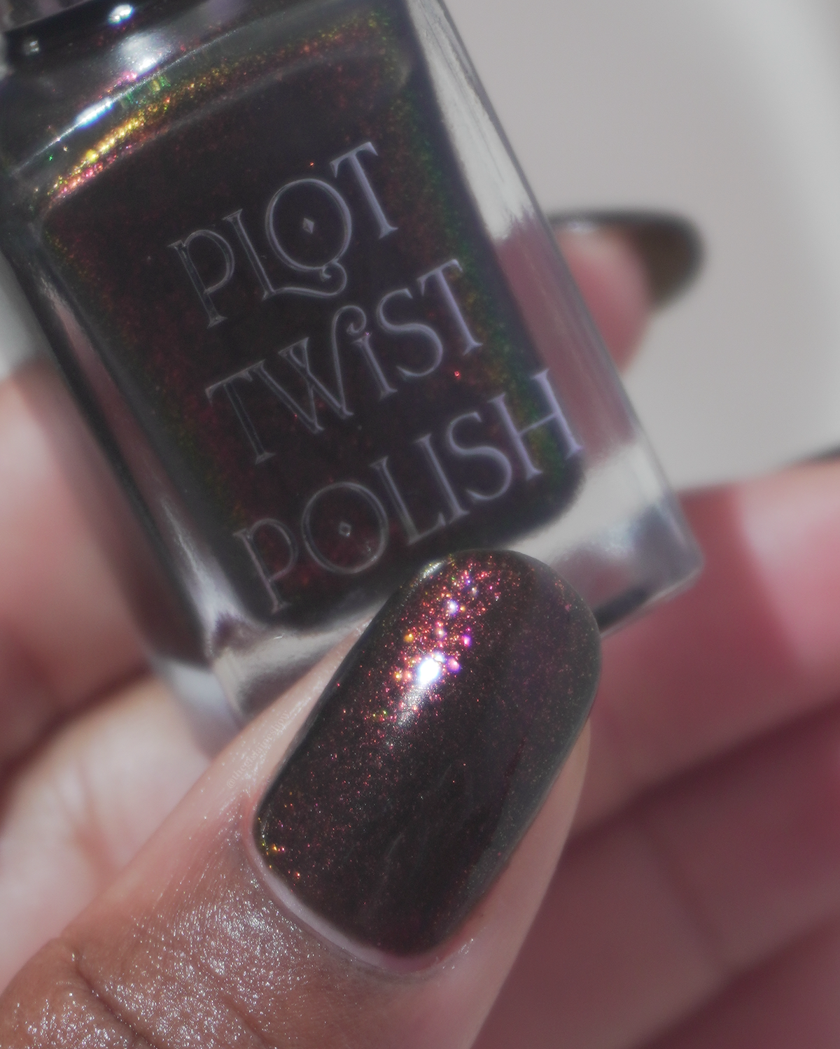 A close-up of a hand holding a bottle of Plot Twist Polish nail lacquer, showing the dark iridescent glitter polish applied to the fingernail.