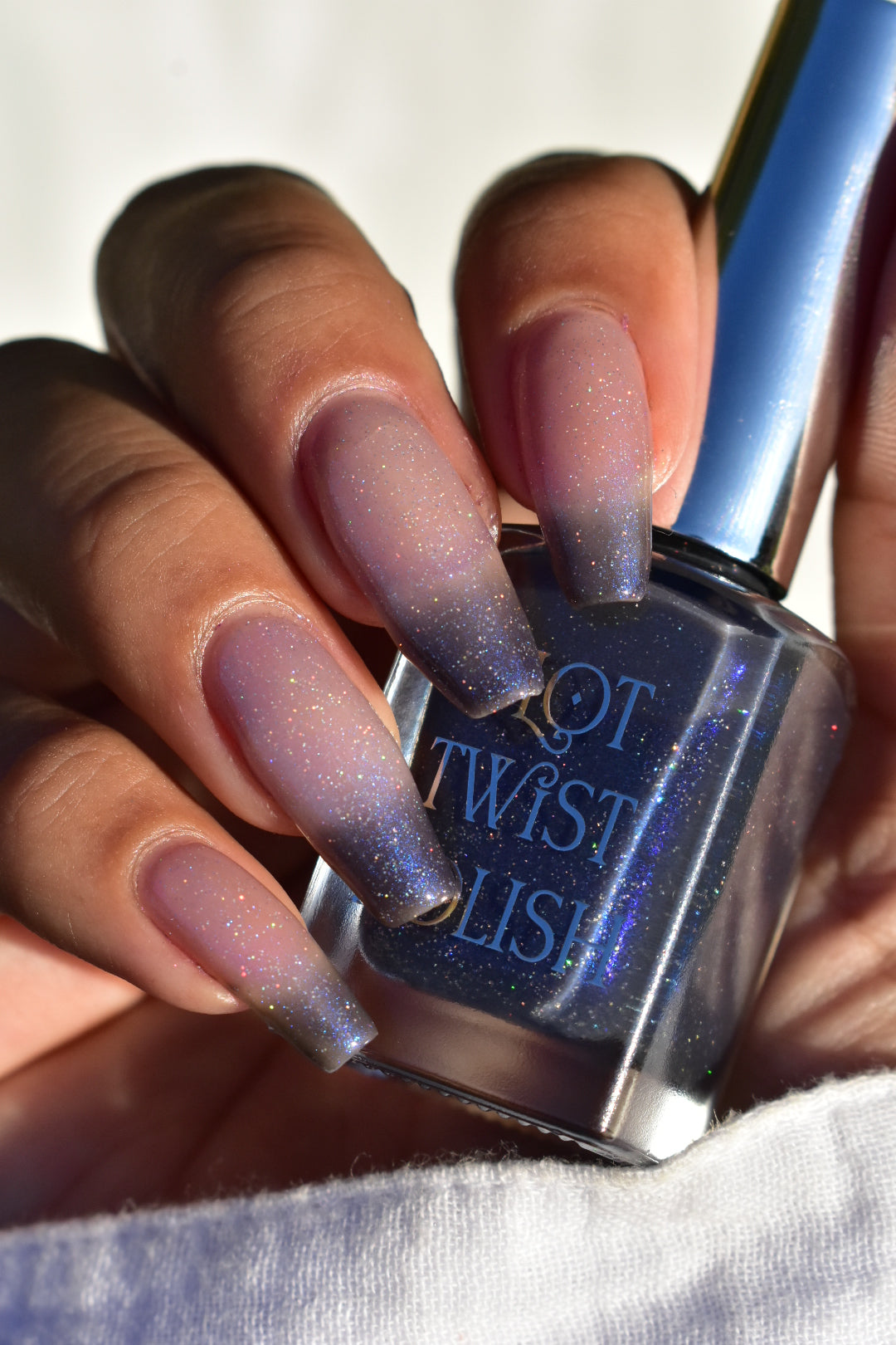 A manicured hand with a translucent shimmery nail polish base and blue nail tips holding a bottle of polish.