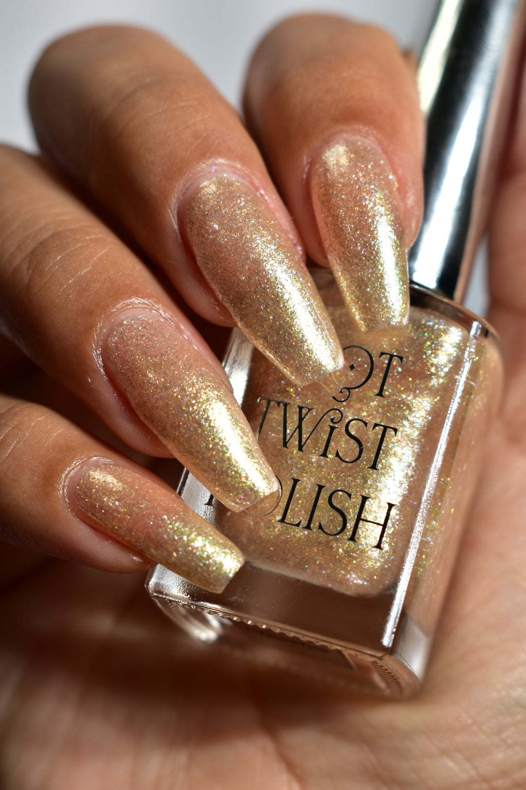 A manicured hand wearing a champagne gold nail polish holding a nail polish bottle. 
