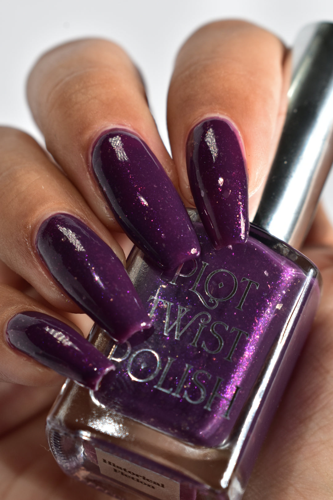 A manicured hand with a dark purple polish with purple shimmer and small gold flakes holding a bottle of nail polish