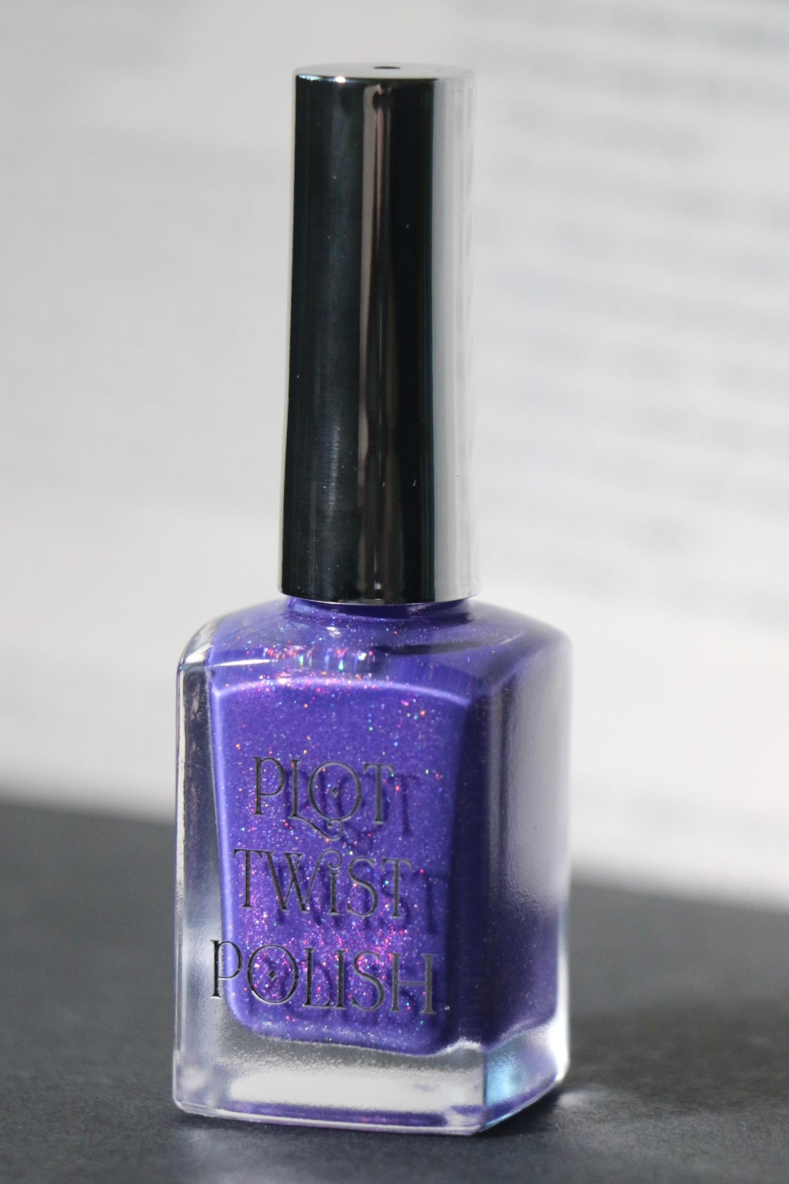 A bottle of Plot Twist Polish nail lacquer in a shimmering purple shade, featuring a silver cap and silver lettering on the clear glass bottle.
