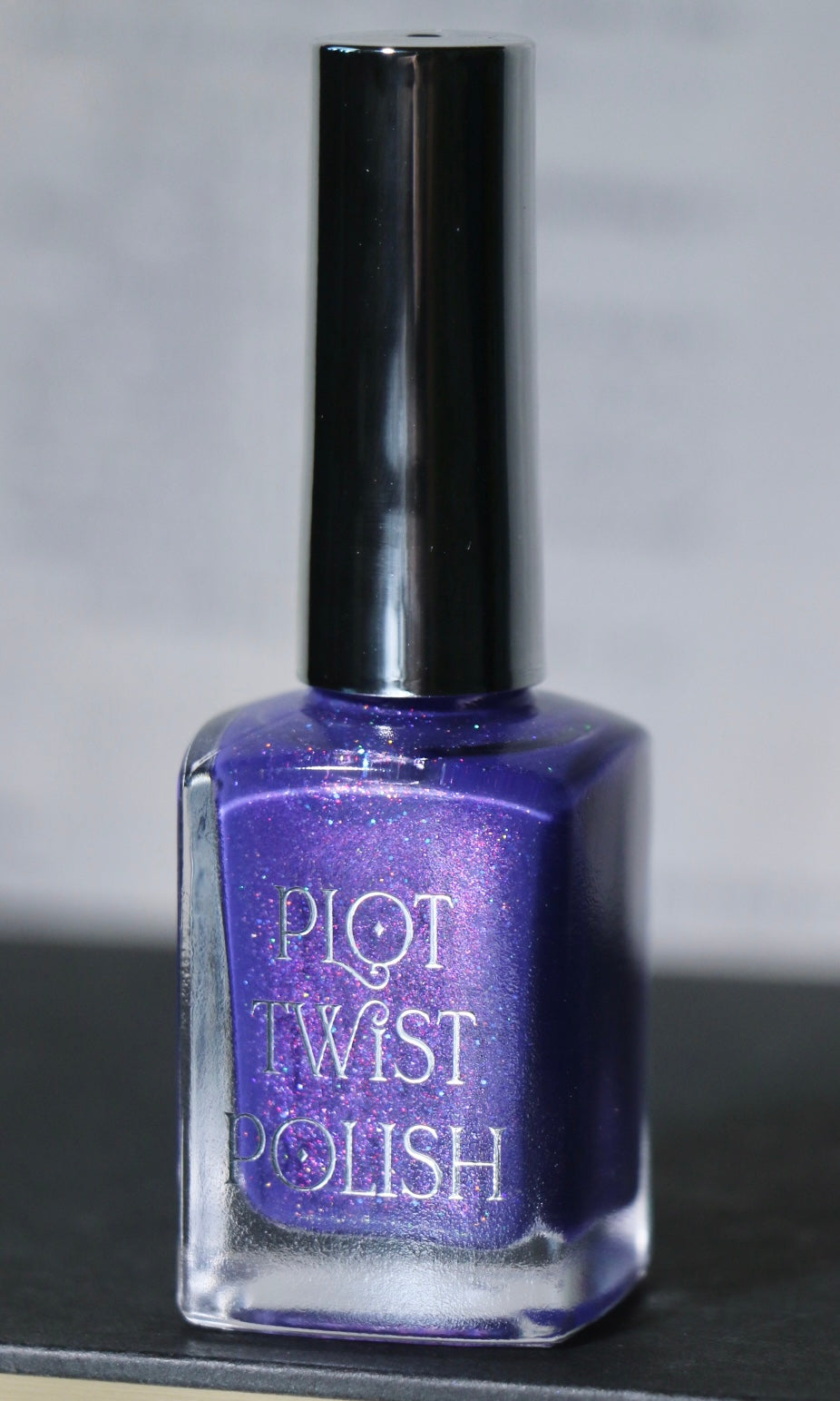 A bottle of Plot Twist Polish nail lacquer in a shimmering purple shade, featuring a silver cap and silver lettering on the clear glass bottle.