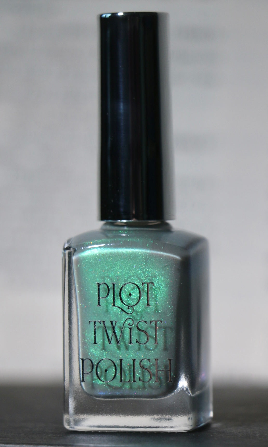 A bottle of Plot Twist Polish nail lacquer in a shimmering grey green green shade, featuring a silver cap and clear glass bottle.
