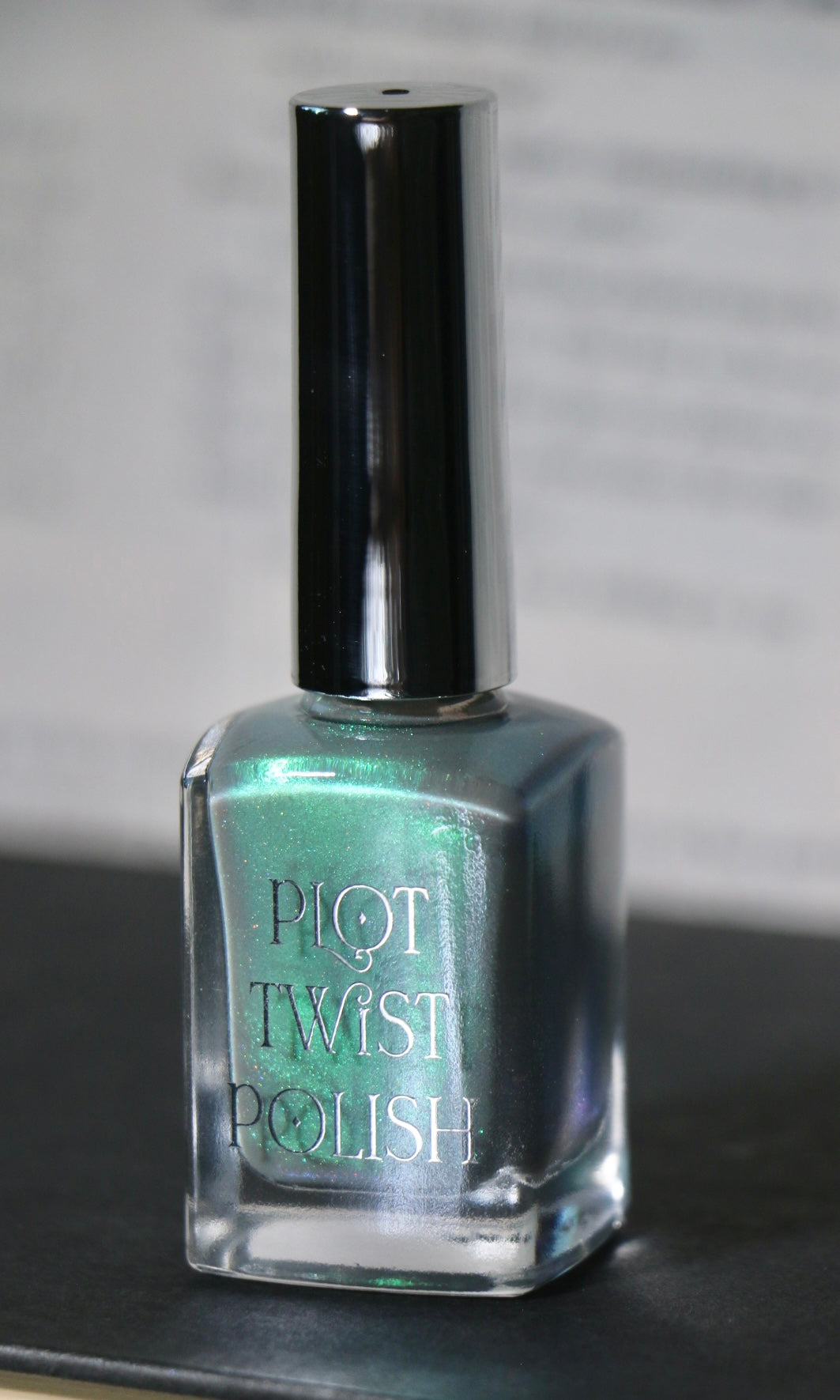 A bottle of Plot Twist Polish nail lacquer in a shimmering grey green shade, featuring a silver cap and clear glass bottle.