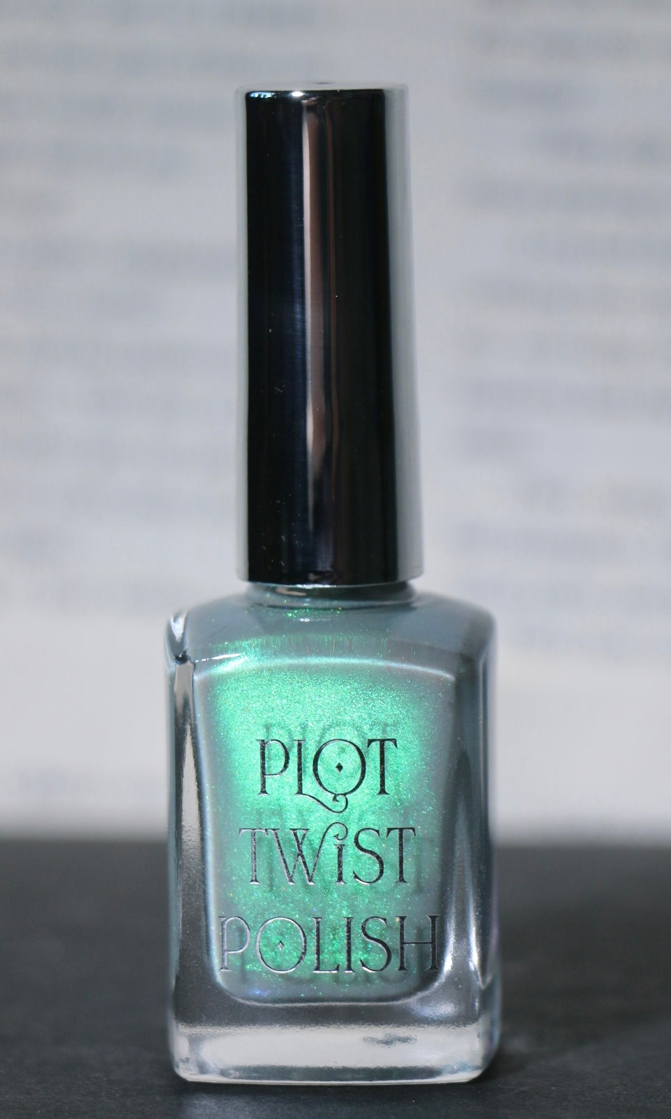 A bottle of Plot Twist Polish nail lacquer in a shimmering grey green green shade, featuring a silver cap and clear glass bottle.