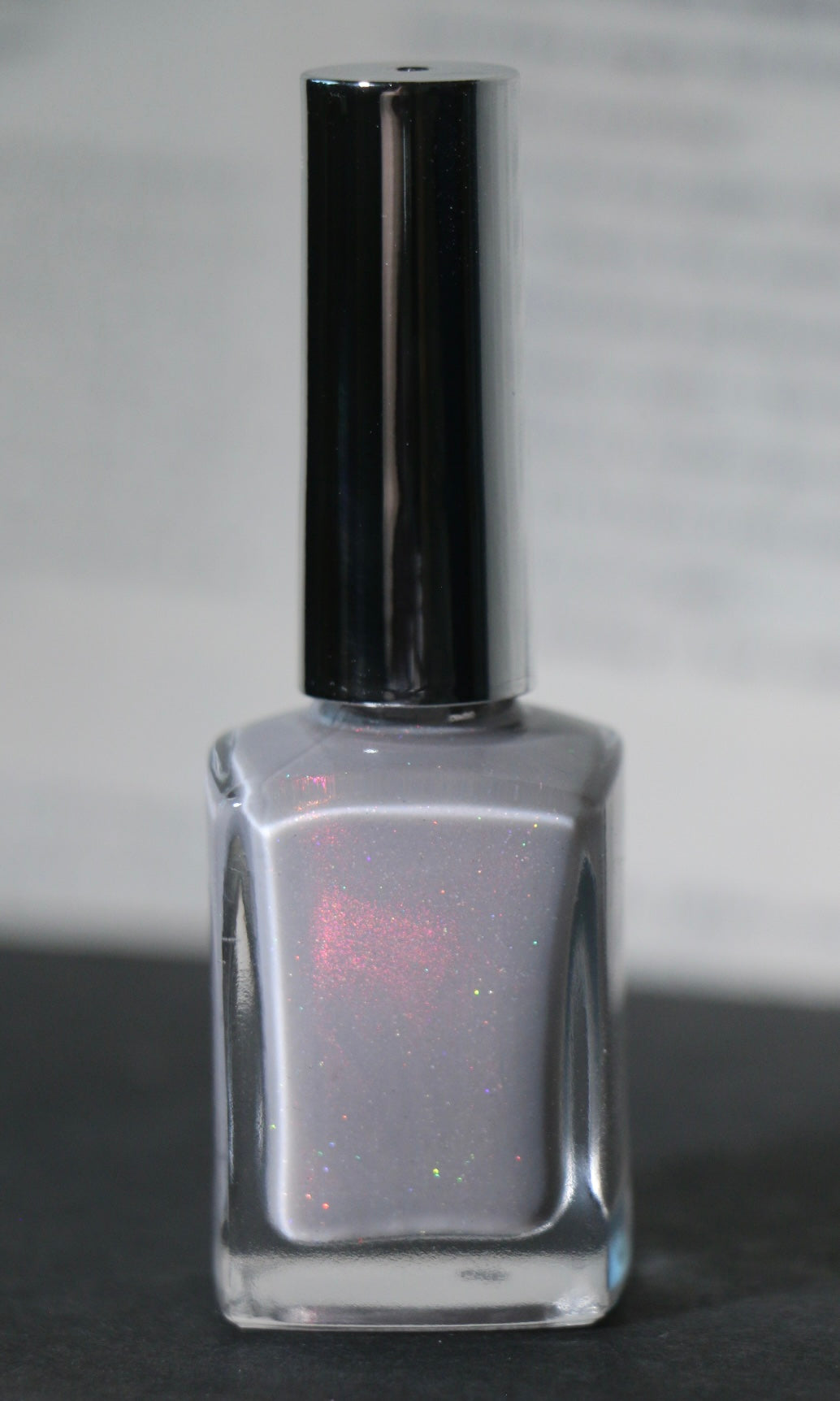 A square glass bottle of nail polish with a reflective silver cap, filled with a shimmering light gray polish that has subtle pink and silver glitter.