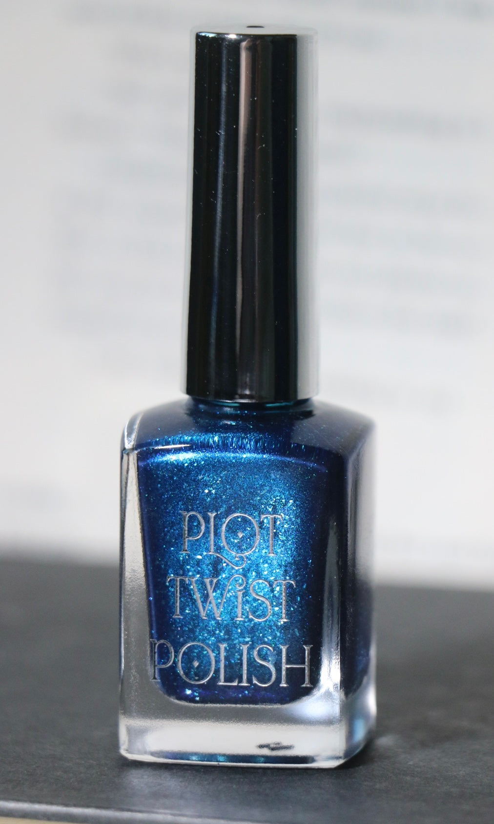 A bottle of Plot Twist Polish nail lacquer in a shimmering metallic blue shade, featuring a silver cap and clear glass bottle, set against a neutral gray background.