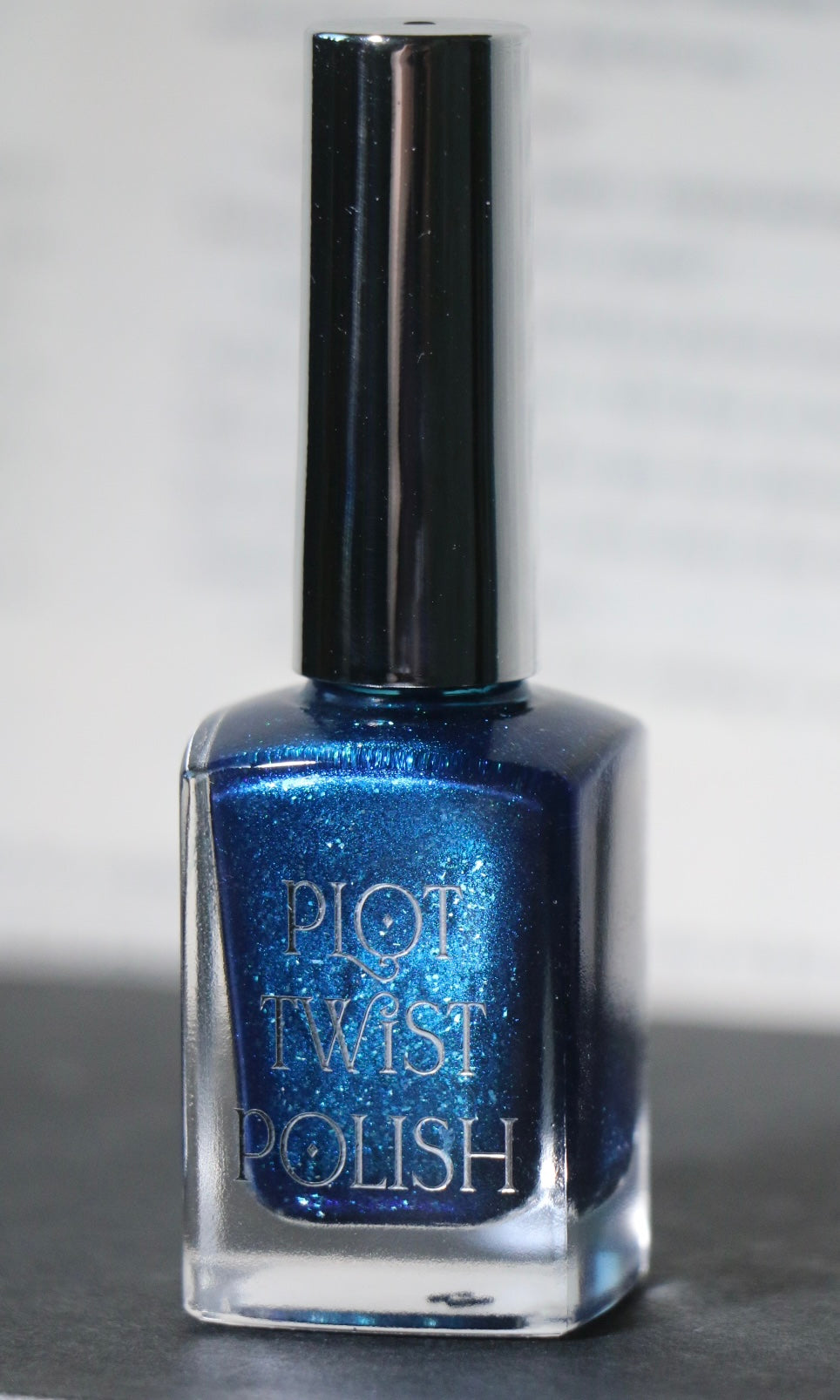 A bottle of blue glitter nail polish with a silver cap, labeled 'PLOT TWIST POLISH', set against a neutral gray background.