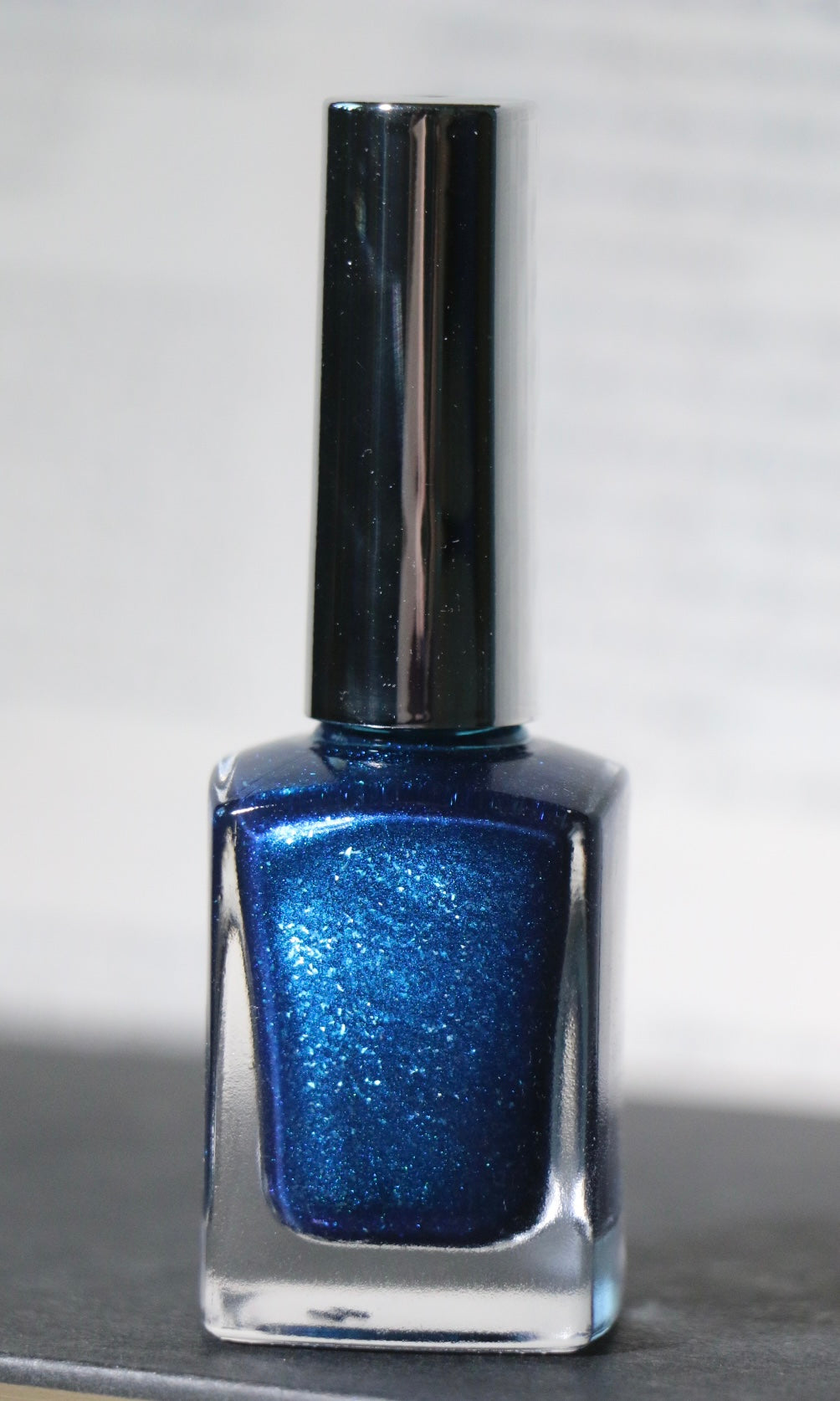 A bottle of shimmering metallic blue nail polish with a reflective silver cap, set against a neutral gray background.