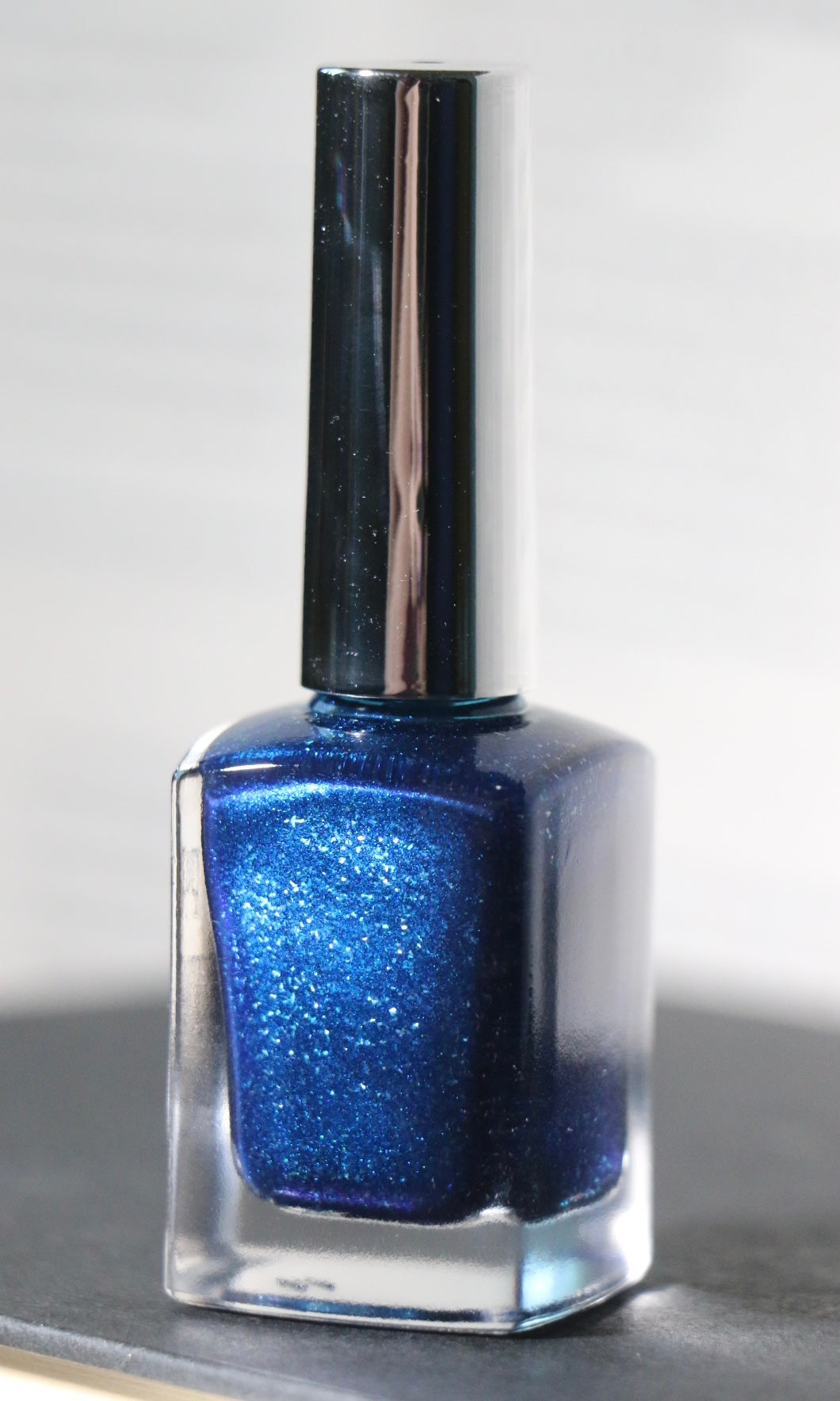 A bottle of blue glitter nail polish with a shiny silver cap, set against a neutral gray background.