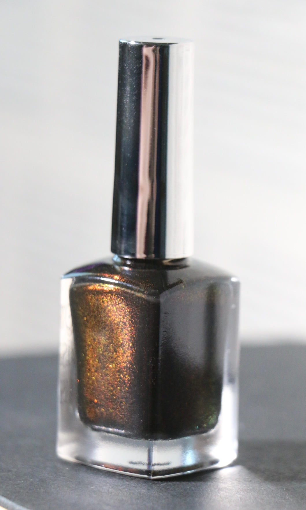 A bottle of metallic dark brown nail polish with shimmering gold and bronze flecks, featuring a reflective silver cap, set against a neutral gray background.