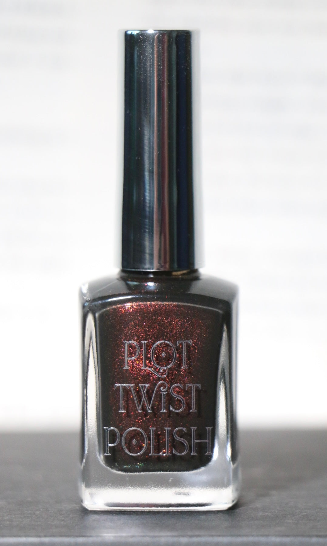 A bottle of Plot Twist Polish nail lacquer in a dark, shimmering metallic bronze-brown shade, featuring a reflective silver cap and clear glass bottle.
