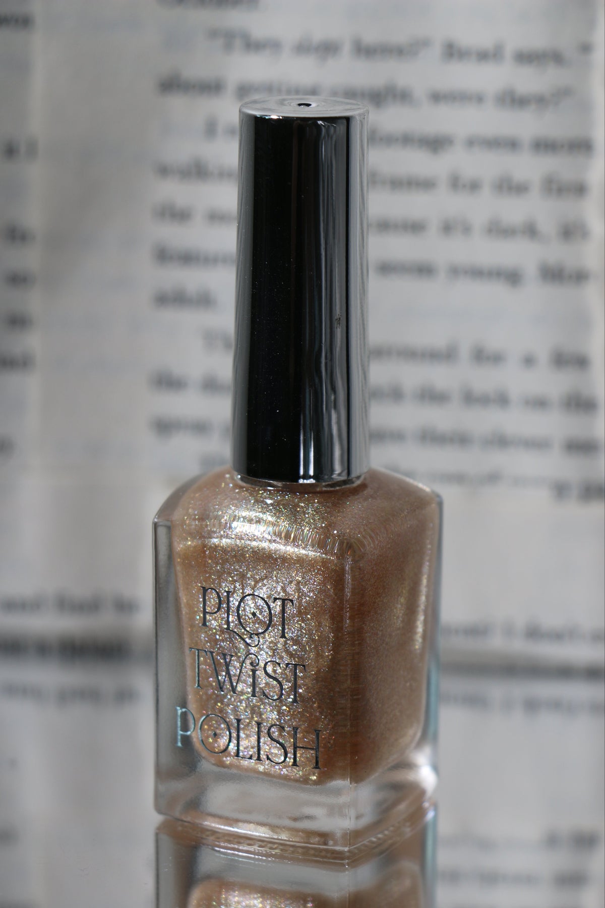 A bottle of Plot Twist Polish nail polish with a shimmering champagne gold nail polish and a glossy silver cap, set against a blurred background of printed book pages.