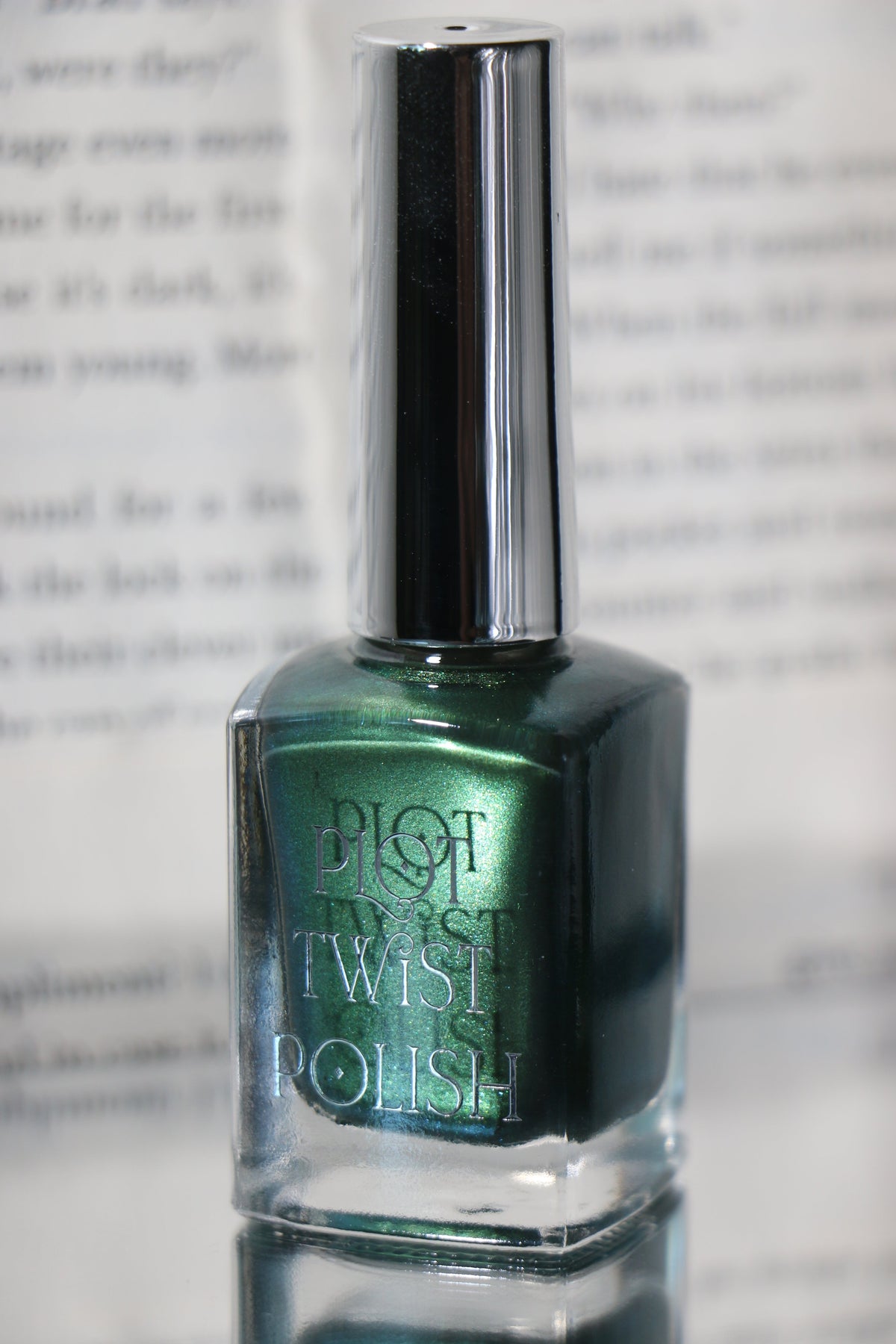 A bottle of Plot Twist Polish nail lacquer in a shimmering metallic emerald green shade, featuring a reflective silver cap, set against a blurred background of printed text.