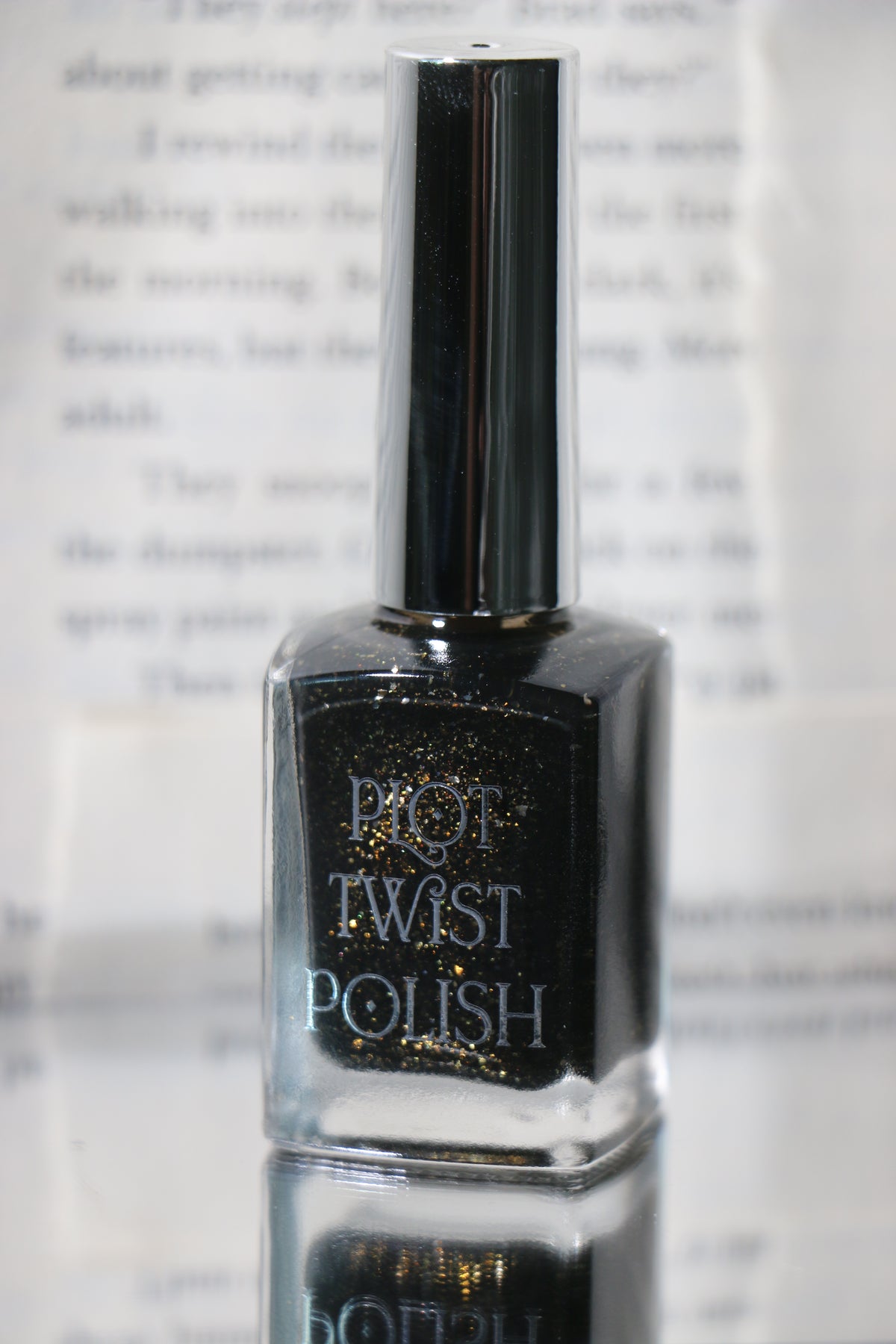 A bottle of Plot Twist Polish nail lacquer in a shimmering black shade with gold glitter, featuring a reflective silver cap, set against a blurred background of printed text.