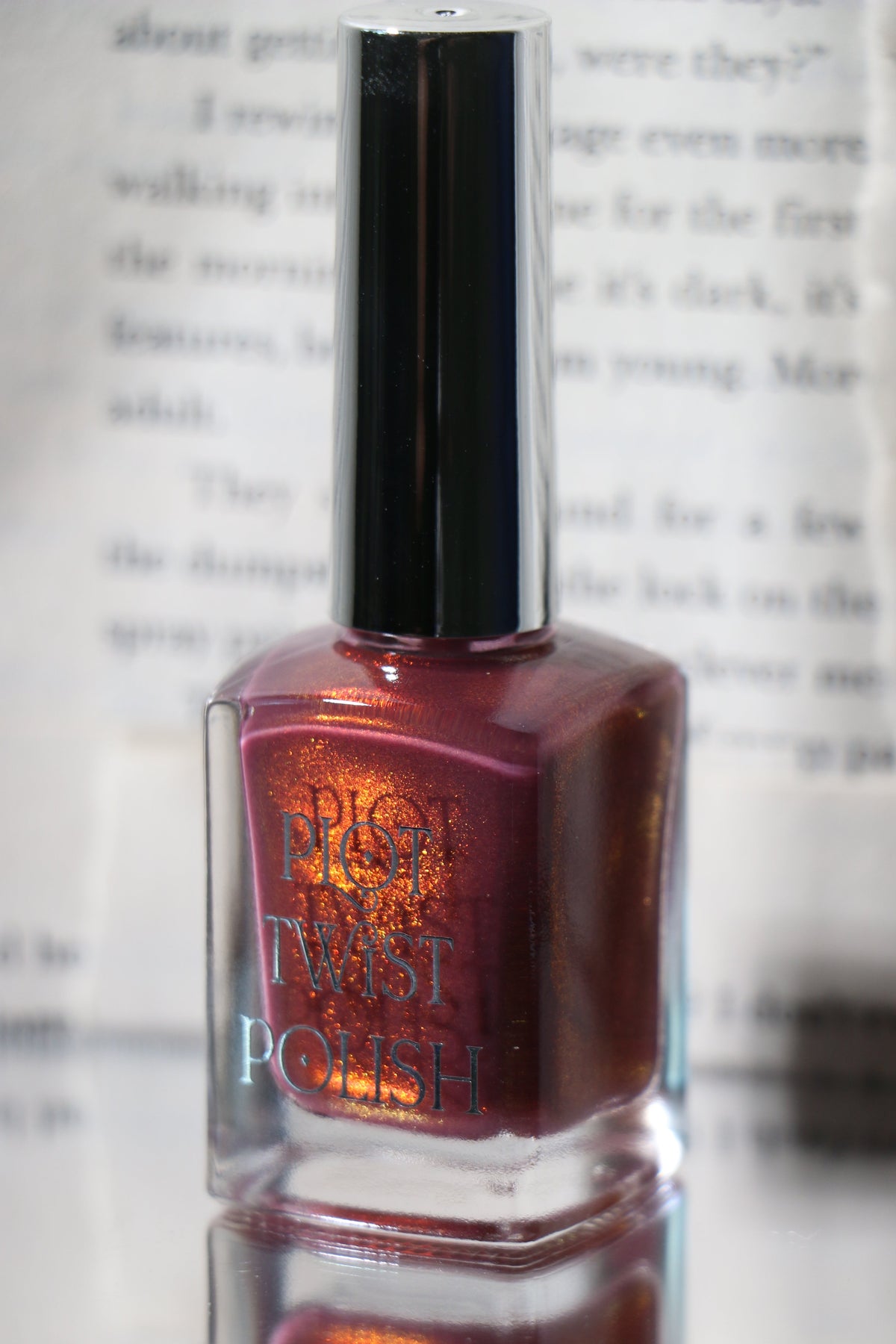 A bottle of Plot Twist Polish nail polish featuring a reflective silver cap, set against a blurred background of printed book text. The nail polish is a rose with a red to orange glow.