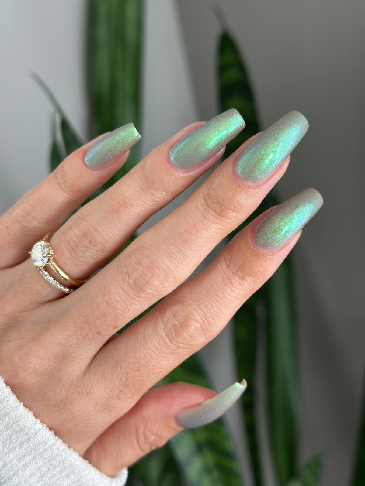 A close-up of a hand with long, almond-shaped nails painted in a shimmering iridescent green and blue manicure. The hand is wearing a gold ring with blurred green leaves visible in the background.