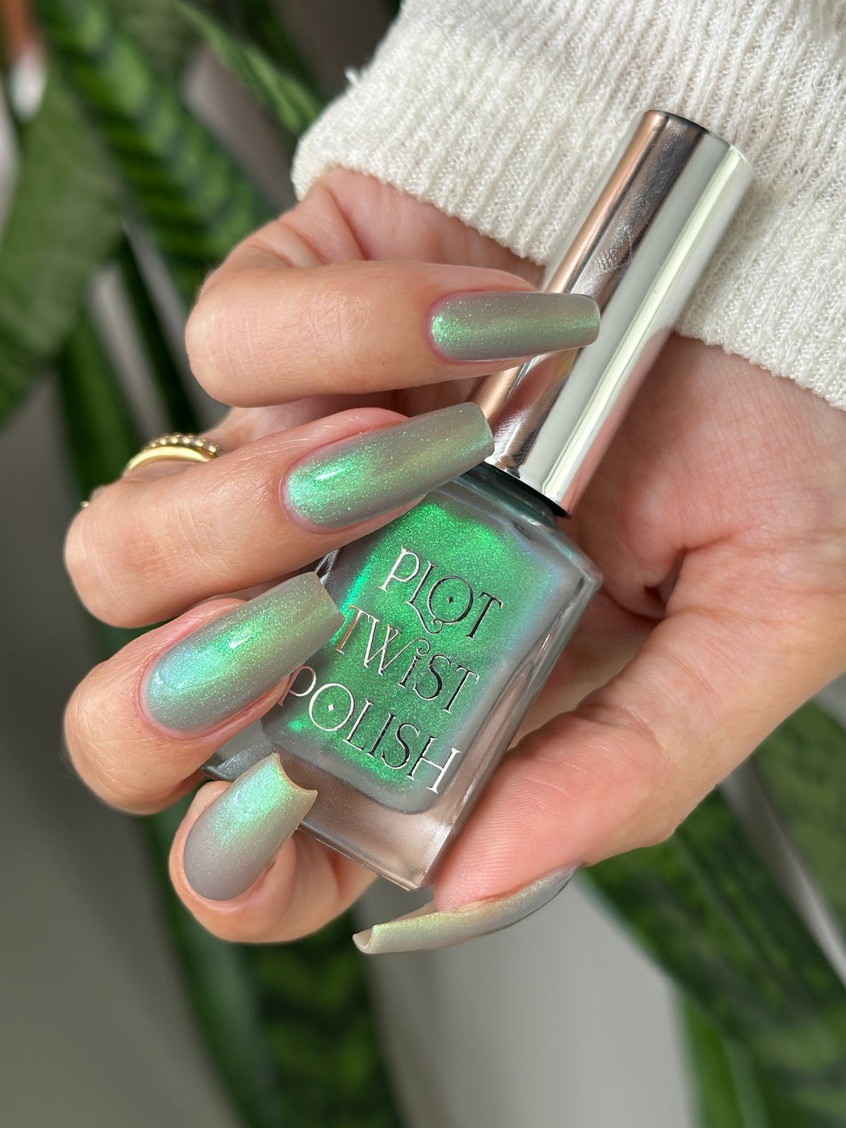 A hand holding a bottle of Plot Twist Polish nail polish, showing off a shimmering iridescent green nail color with a blue shift. The hand is wearing a white knit sweater cuff and a gold ring, with blurred green plants in the background.