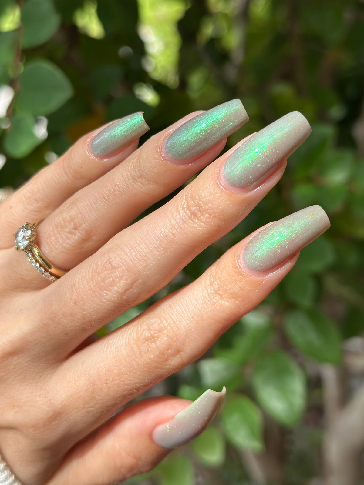 A close-up of a hand with long, coffin-shaped nails painted in a soft green with holographic glitters manicure, with a gold ring on one finger against a blurred green foliage background.