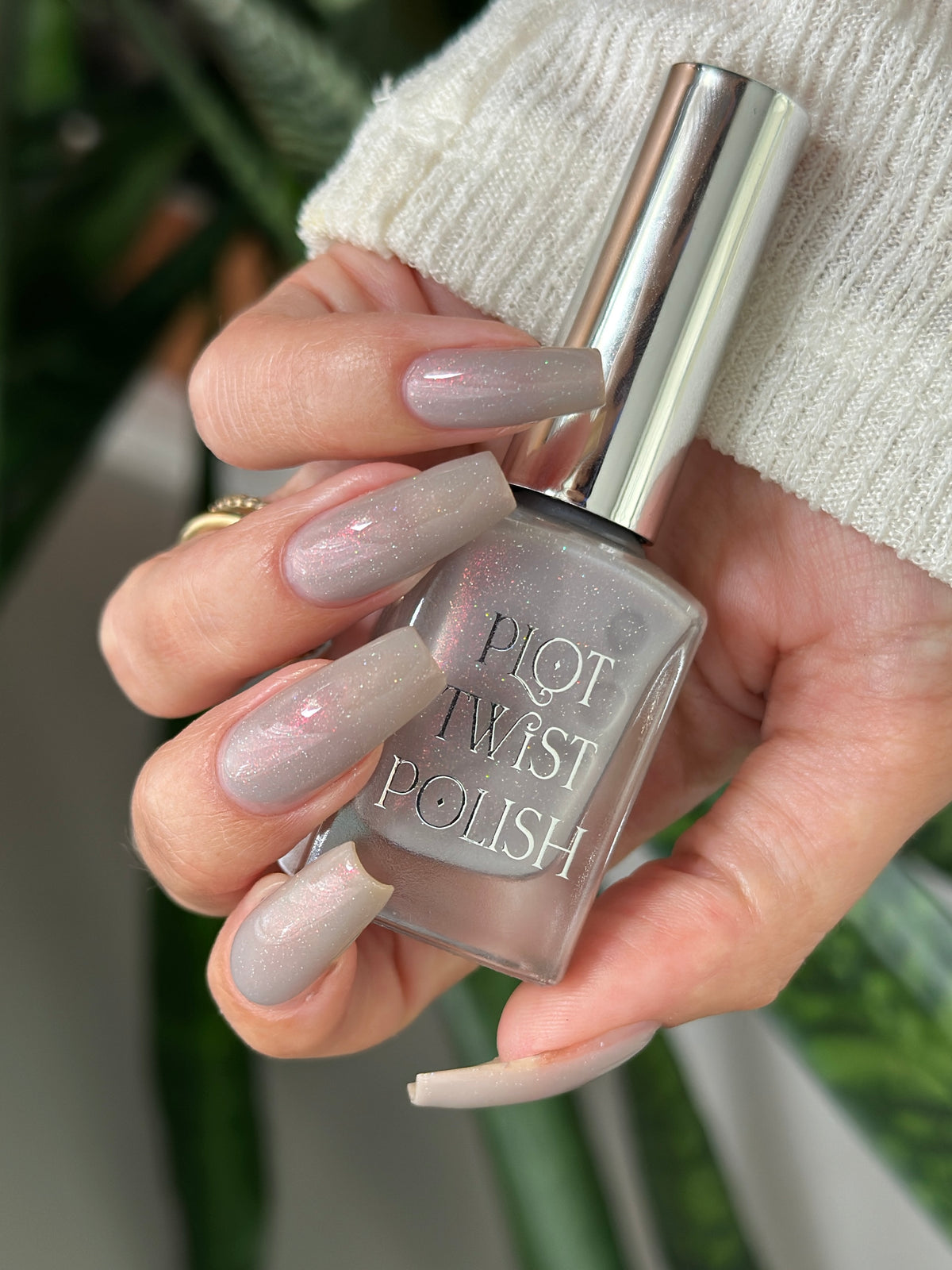 A hand holding a bottle of Plot Twist Polish nail polish, showing off a glossy, light gray manicure. 
