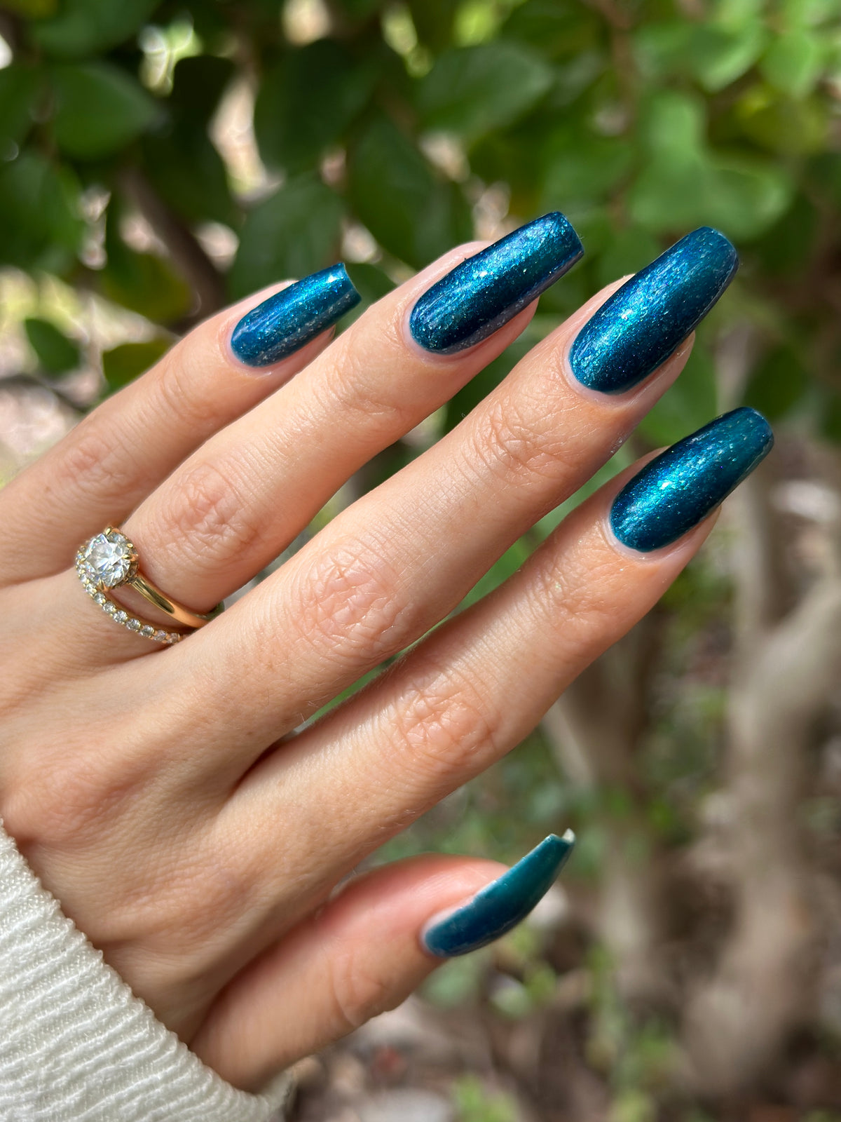 A close-up of a hand with long, almond-shaped nails painted in a shimmering metallic teal-blue nail polish, with a gold ring on one finger and a blurred outdoor background of green leaves.