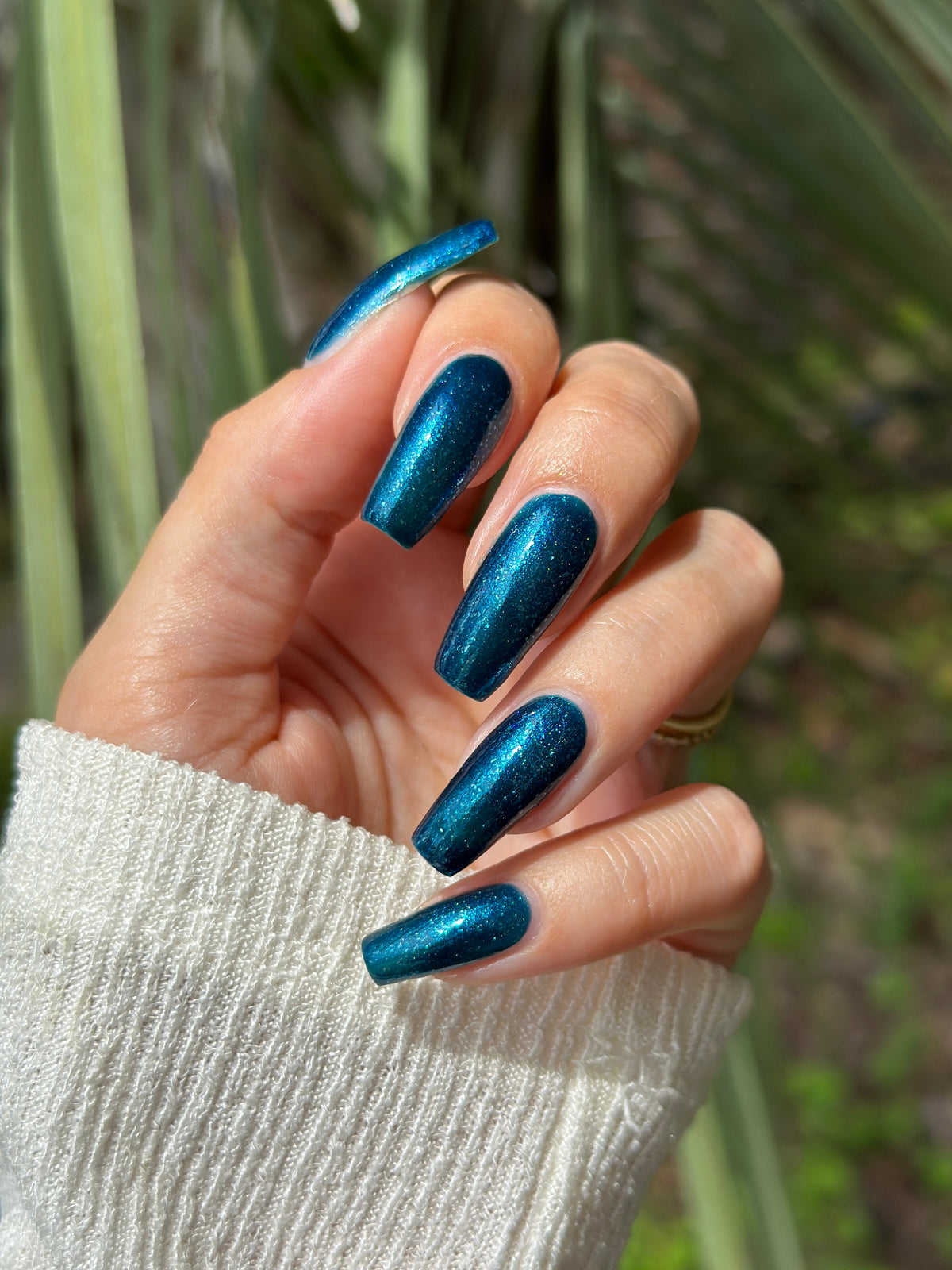 A close-up of a hand with long, almond-shaped nails painted in a shimmering metallic teal-blue nail polish, wearing a white ribbed knit sweater sleeve against a blurred green outdoor background.
