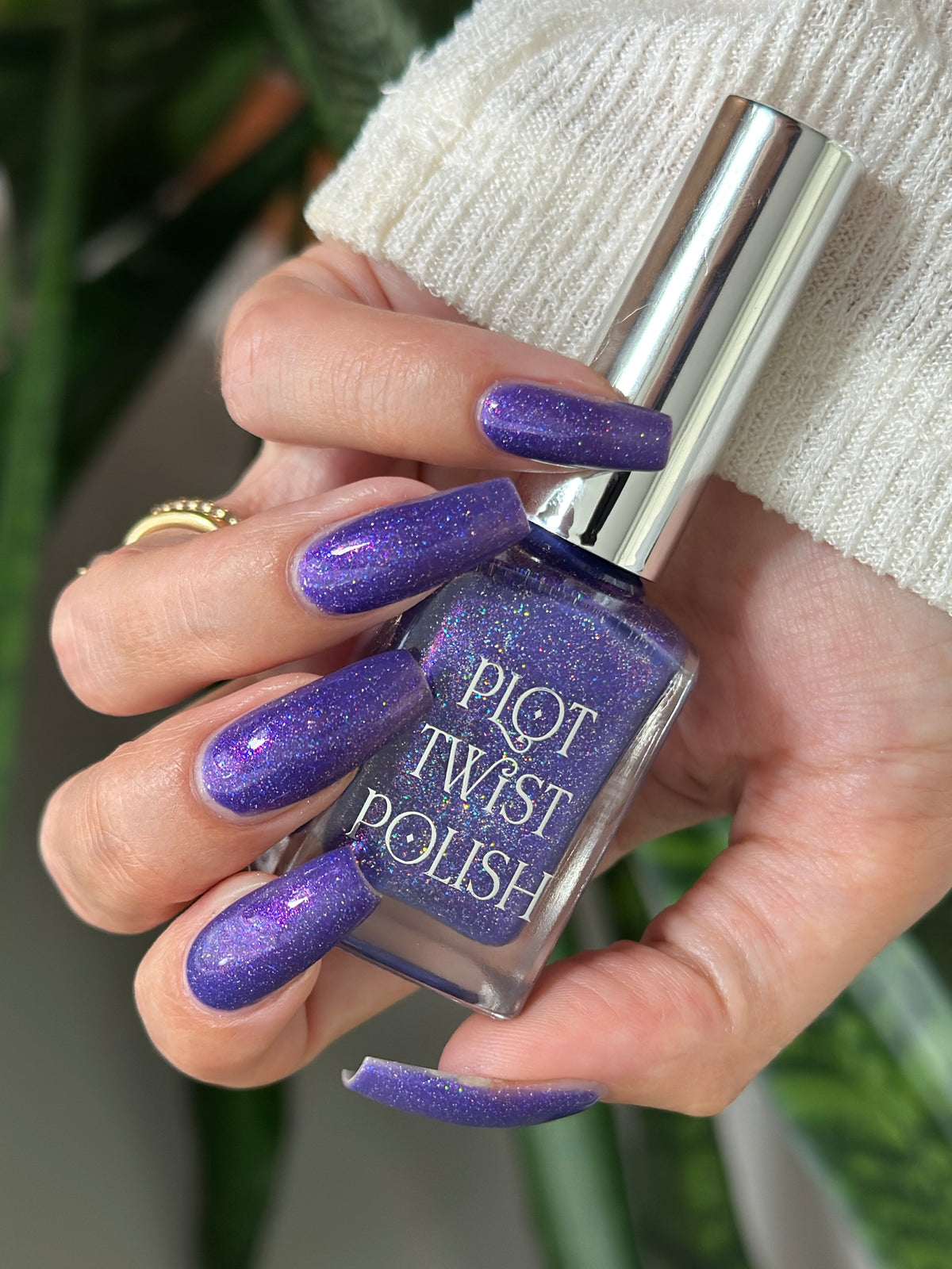 A hand holding a bottle of Plot Twist Polish nail lacquer in a sparkling purple shade, with the nails painted to match. The hand is wearing a white knit sweater and a gold ring, with a blurred green plant in the background.