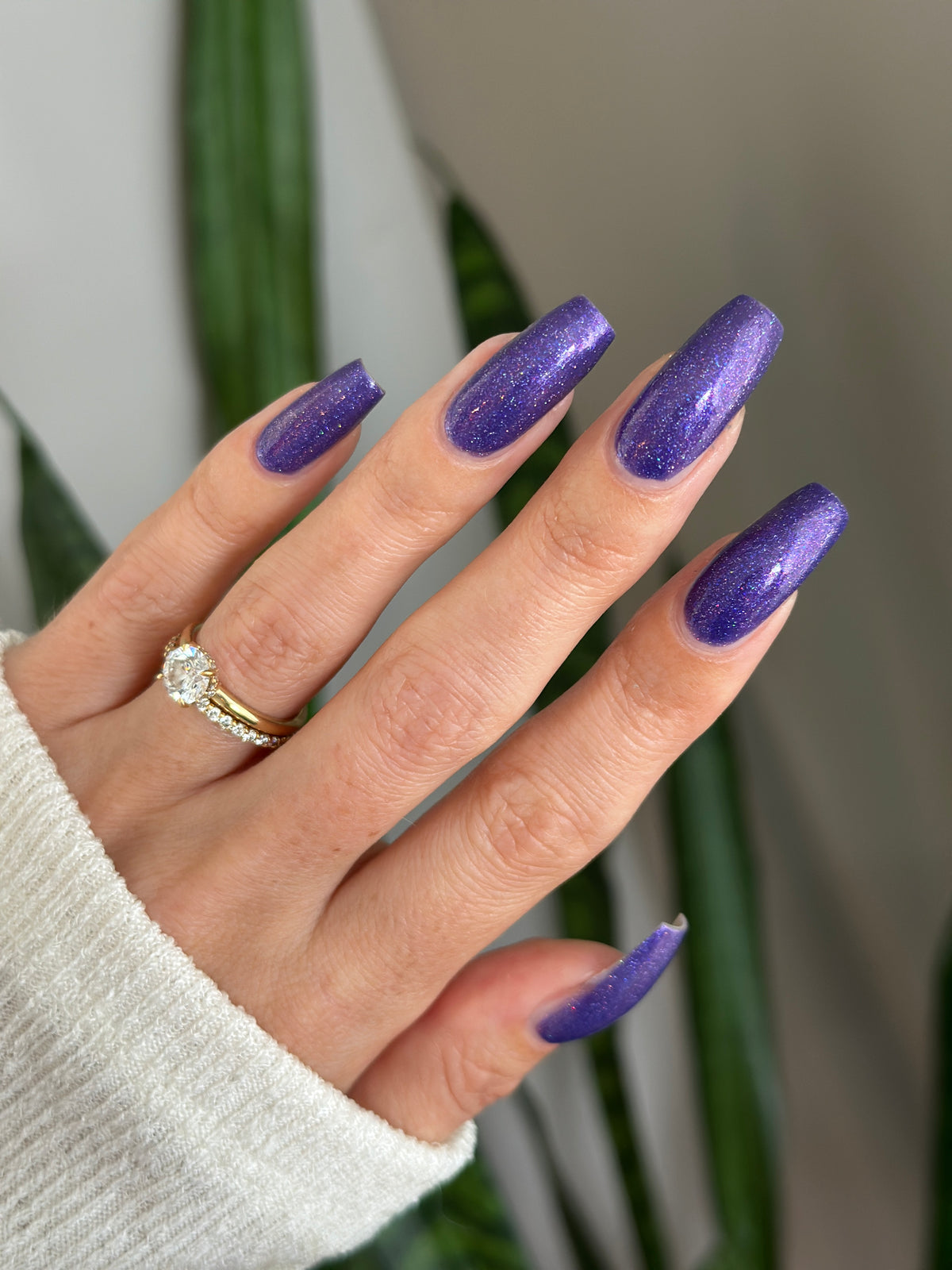 A close-up of a hand with long, almond-shaped nails painted in a sparkling purple glitter polish. 