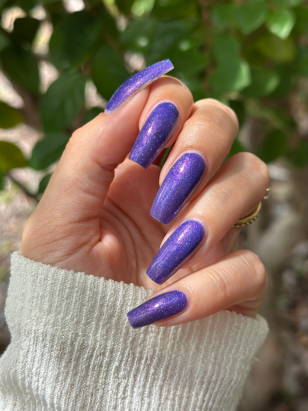 A close-up of a hand with long, almond-shaped nails painted in a sparkling purple glitter nail polish, set against a blurred background of green leaves.