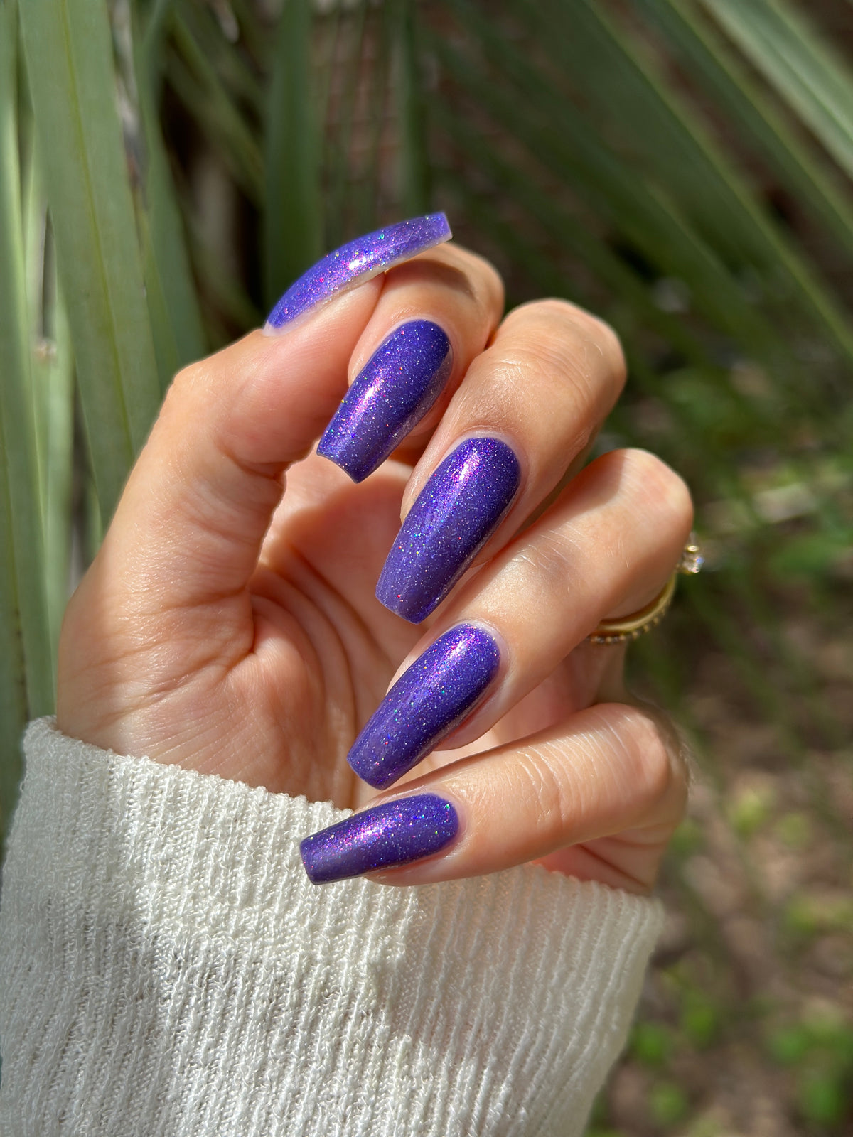 A close-up of a hand with long, stiletto-shaped nails painted in a glittery purple polish, wearing a white ribbed sweater sleeve and a gold ring, with blurred green foliage in the background.