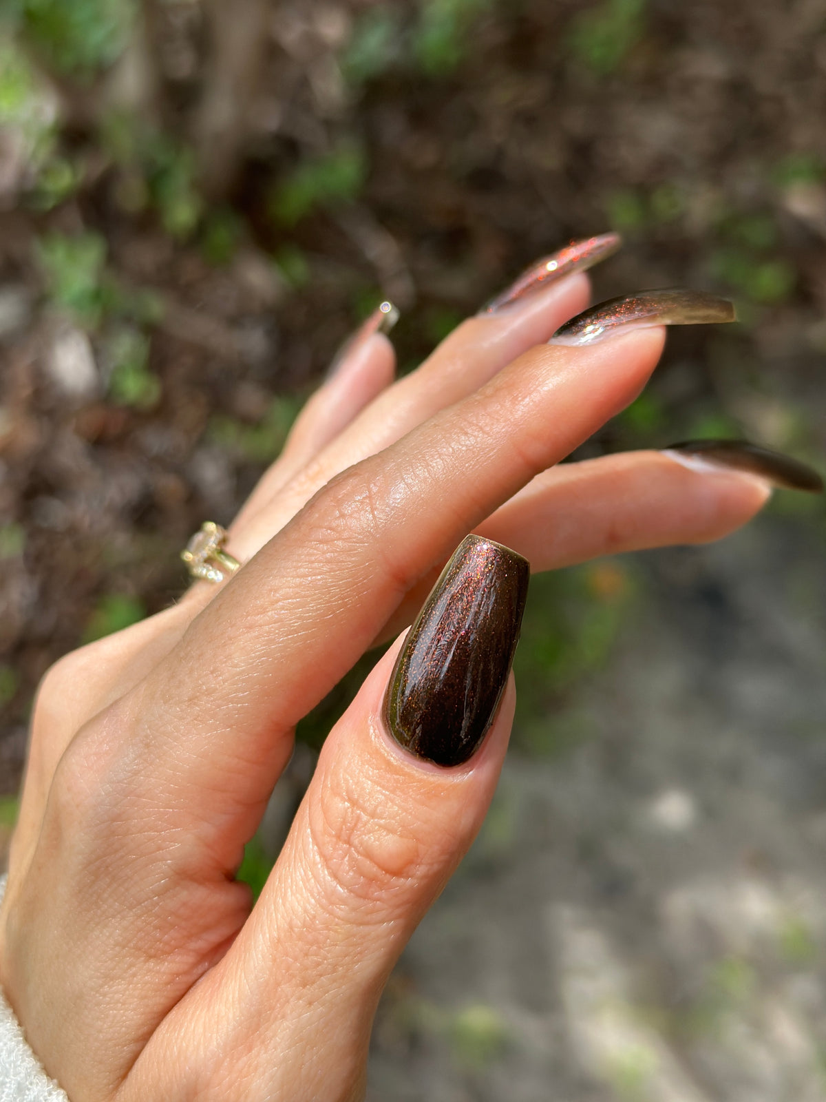 A close-up of a hand with long, glossy dark brown coffin-shaped nails, set against a blurred outdoor background of green foliage and brown ground.