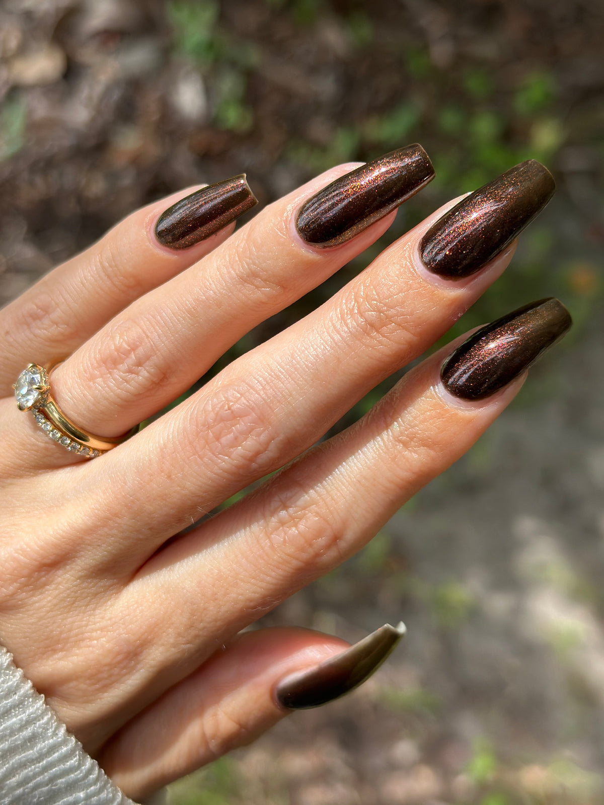 A close-up of a hand with long, almond-shaped nails painted in a metallic dark brown shimmer, with a gold ring on one finger, set against a blurred outdoor background.