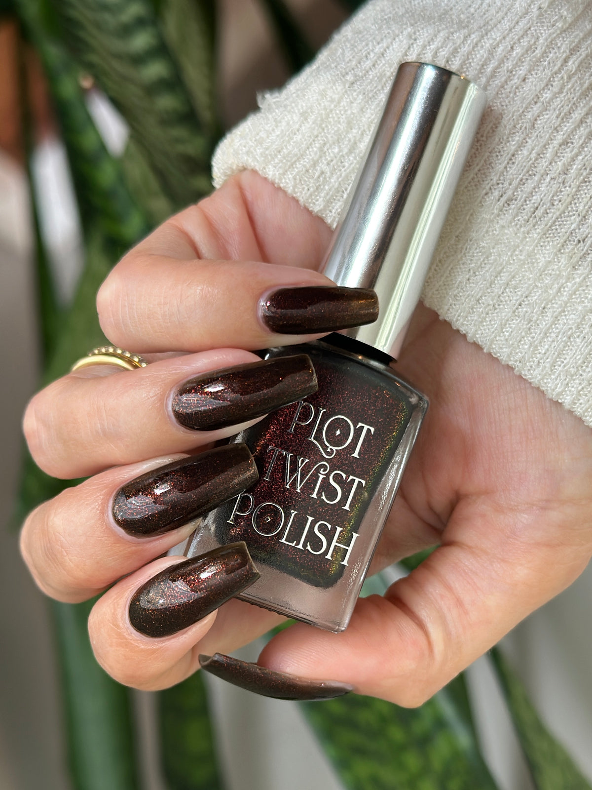 A hand holding a bottle of Plot Twist Polish nail lacquer with a dark metallic brown glitter polish. The hand has the same manicured nails painted in the matching dark brown shade, and is wearing a white knit sweater and a gold ring.