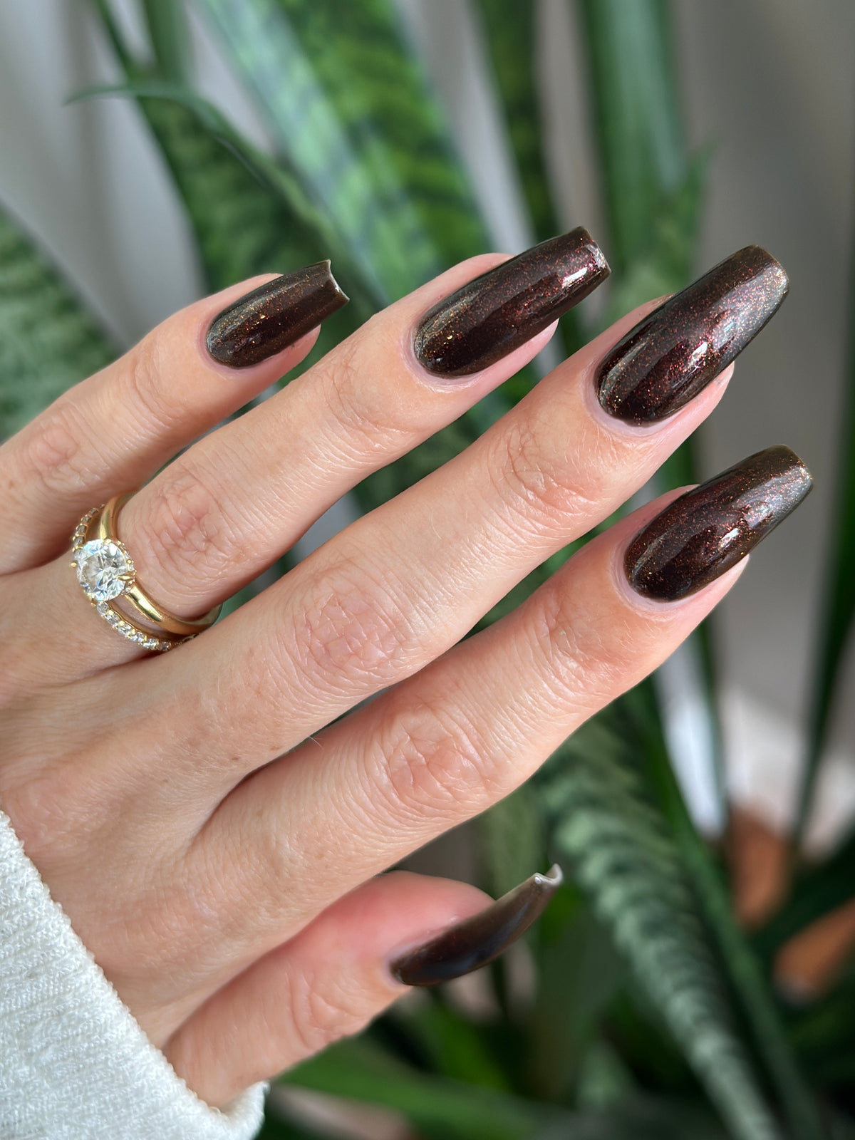 A close-up of a hand with long, glossy dark brown nail polish with subtle shimmer, wearing a gold ring, with a blurred green plant in the background.