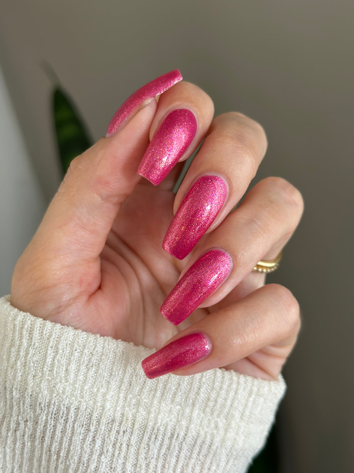 A close-up of a hand with long, almond-shaped nails painted in a sparkling metallic pink nail polish, wearing a white knit sweater and a gold ring.