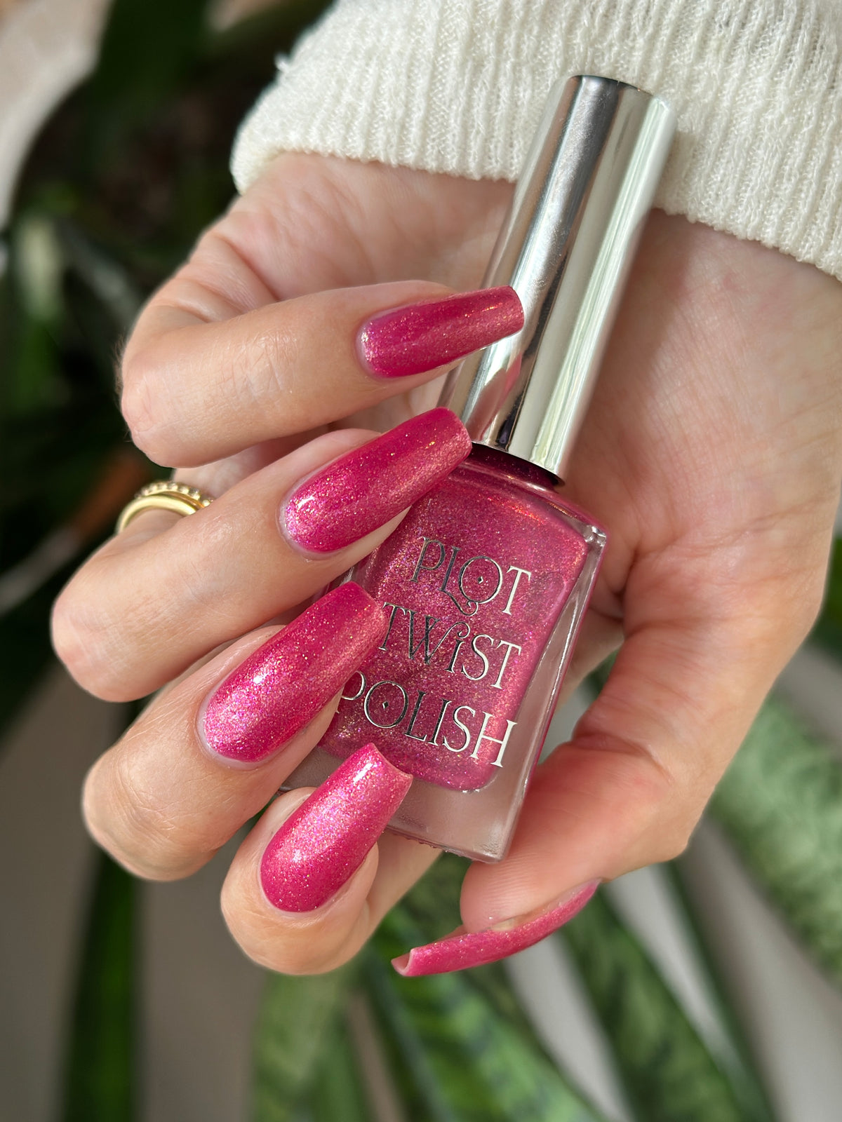 A hand holding a bottle of hot pink glitter nail polish, with the nails painted in the same shade. The bottle has a silver cap and the text 'PLOT TWIST POLISH' is visible on the front.