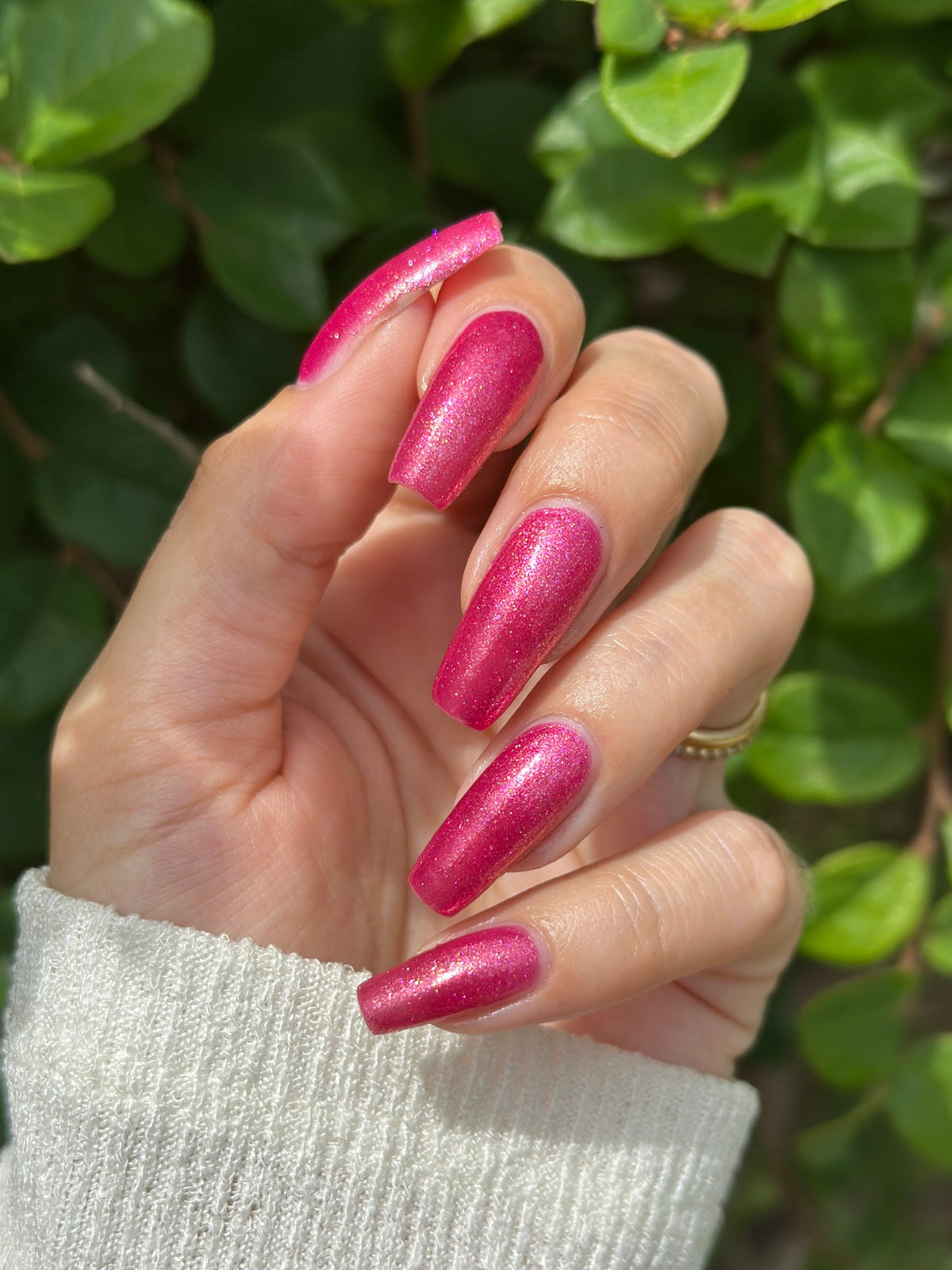 A close-up of a hand with long, almond-shaped nails painted in a sparkling metallic pink polish, set against a background of green leaves.