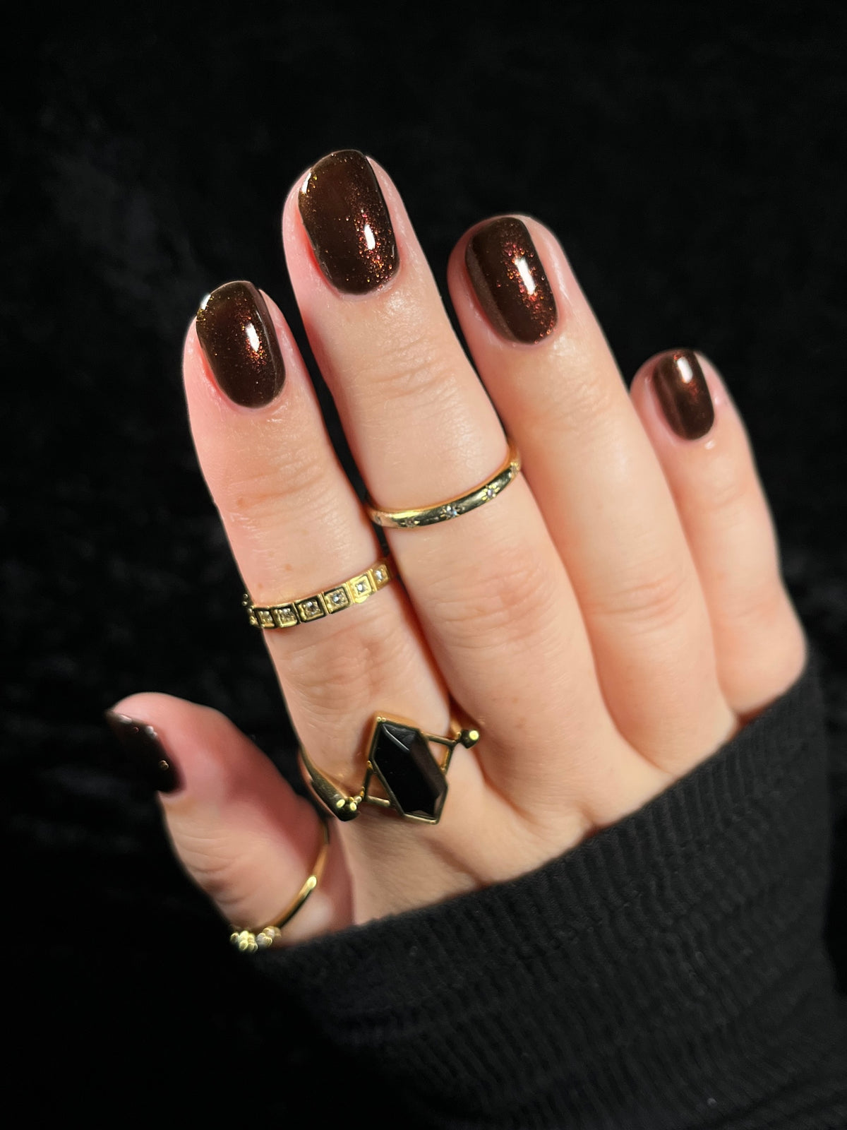 A close-up of a hand wearing multiple gold rings with brown painted nails against a black background.