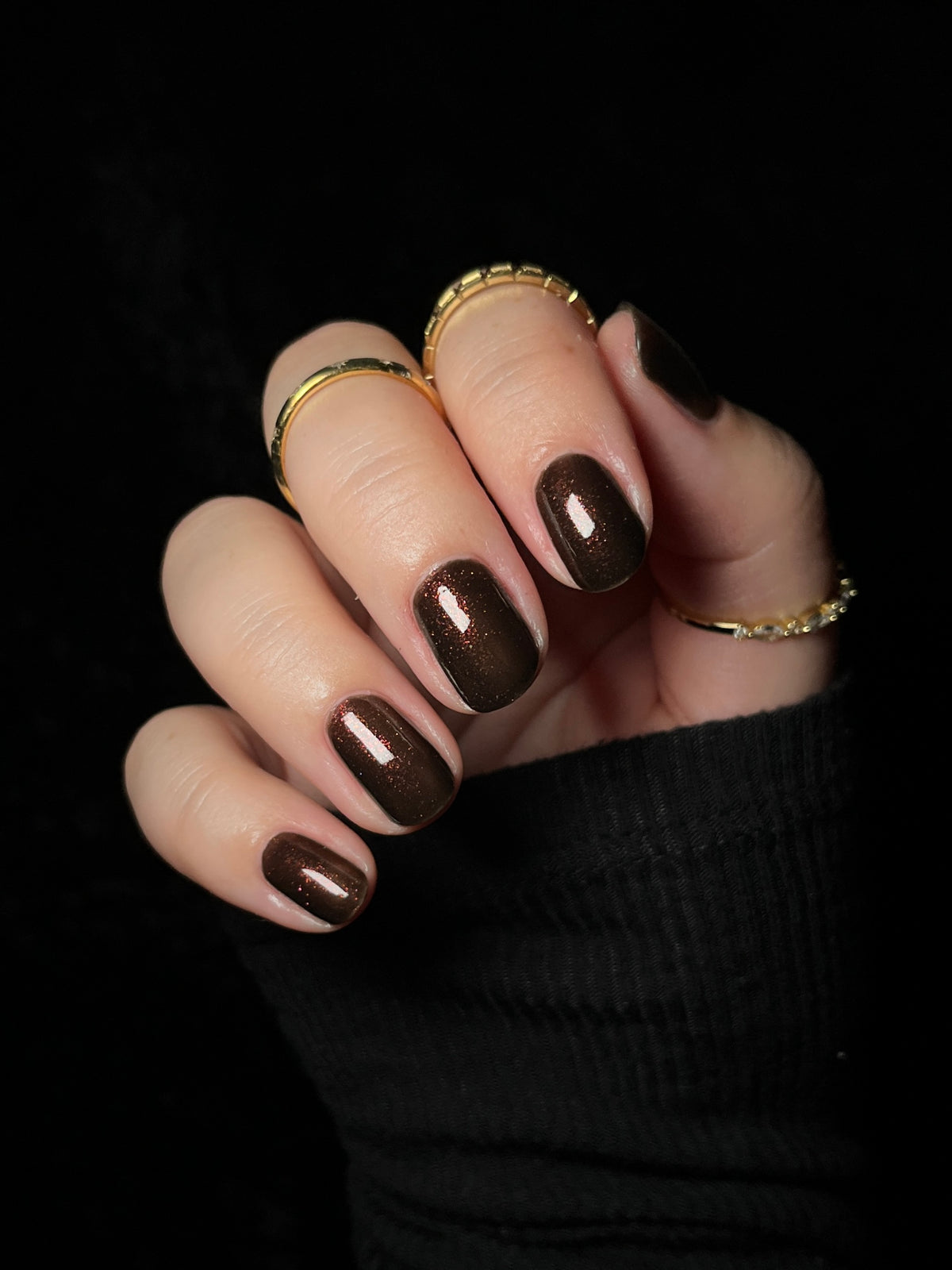 A close-up of a hand with manicured nails painted in a glossy dark brown color with fine gold glitter, wearing several gold rings against a solid black background.