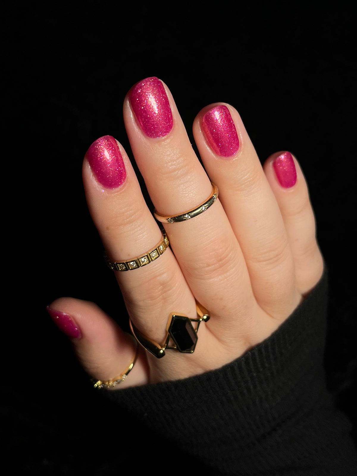 A close-up of a hand with glittery magenta pink nail polish, wearing multiple gold rings, set against a solid black background.