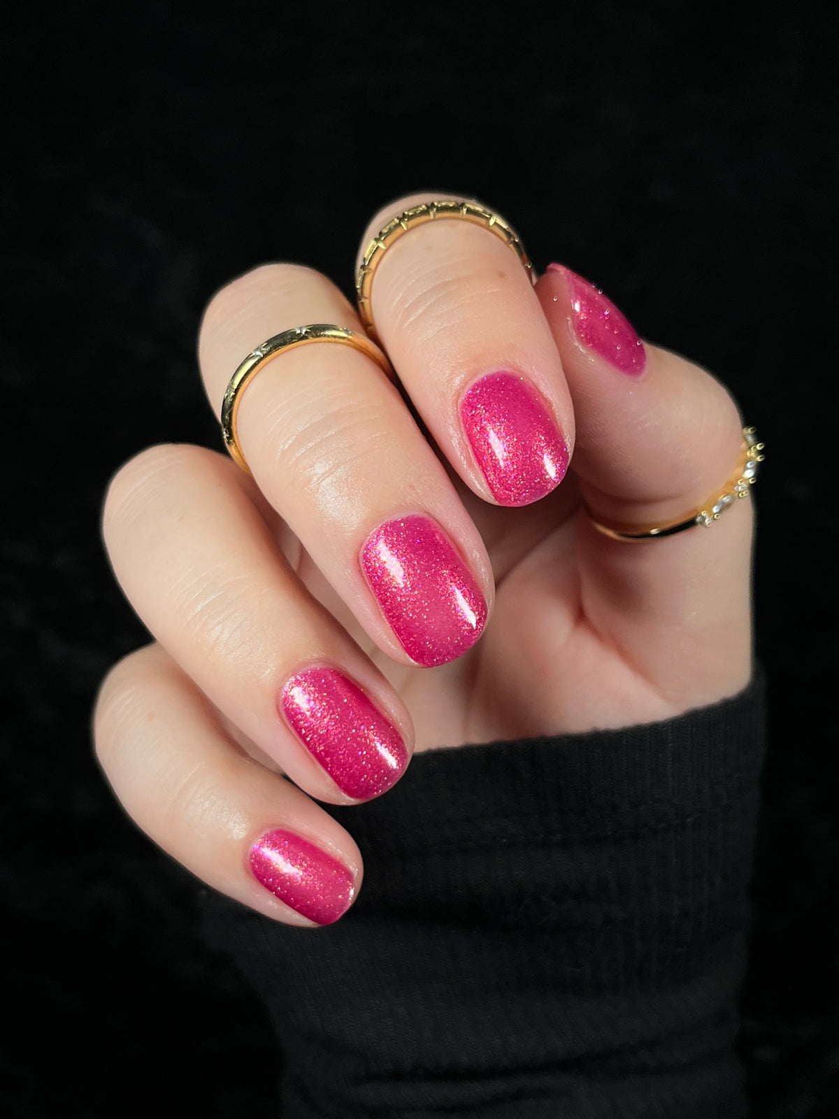A close-up of a hand with manicured nails painted in a sparkling medium pink polish, wearing several gold rings against a black background.