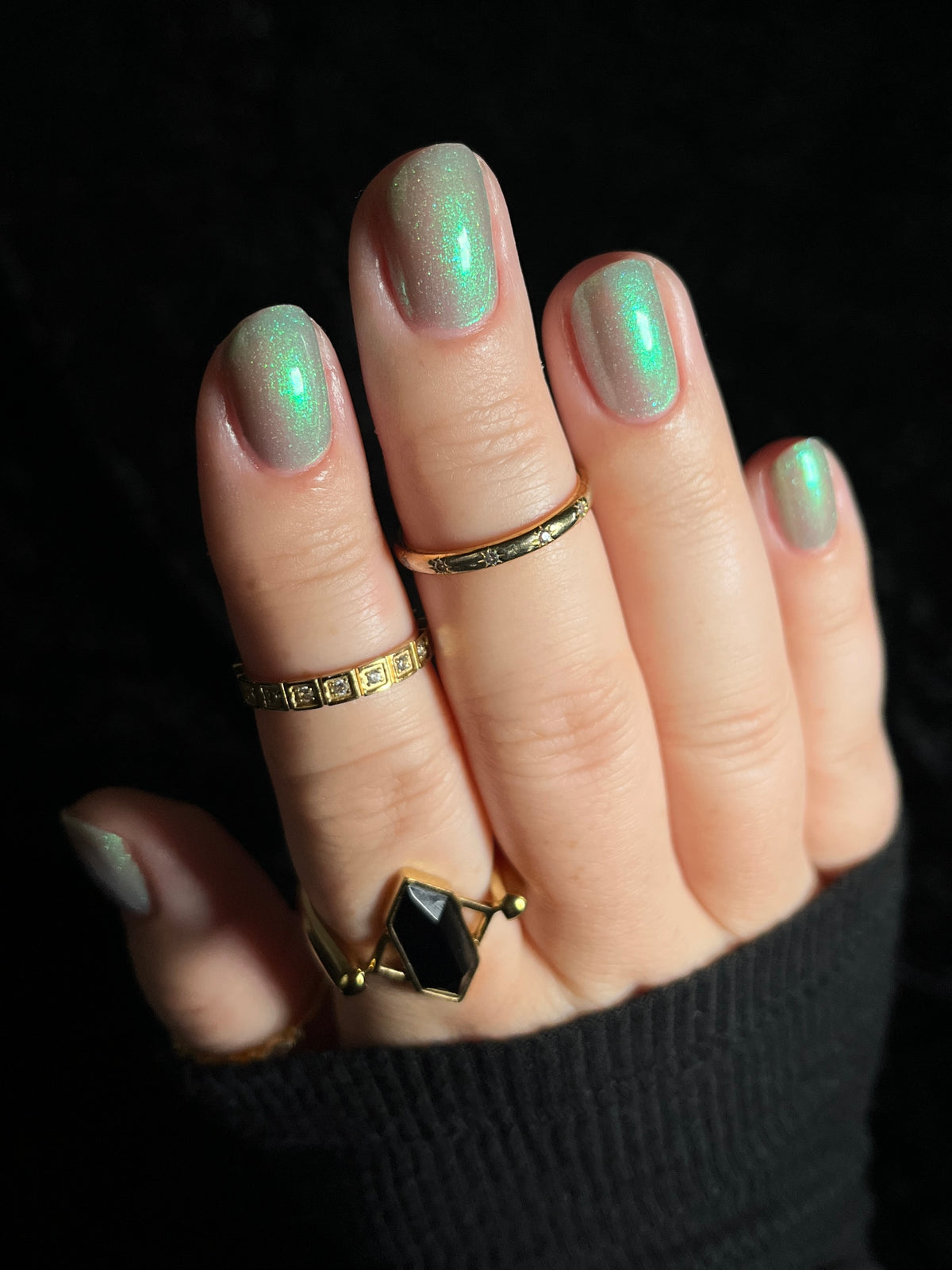 A close-up of a hand with manicured nails painted in a shimmering green color, wearing several gold rings, including a geometric black stone ring, set against a dark background.