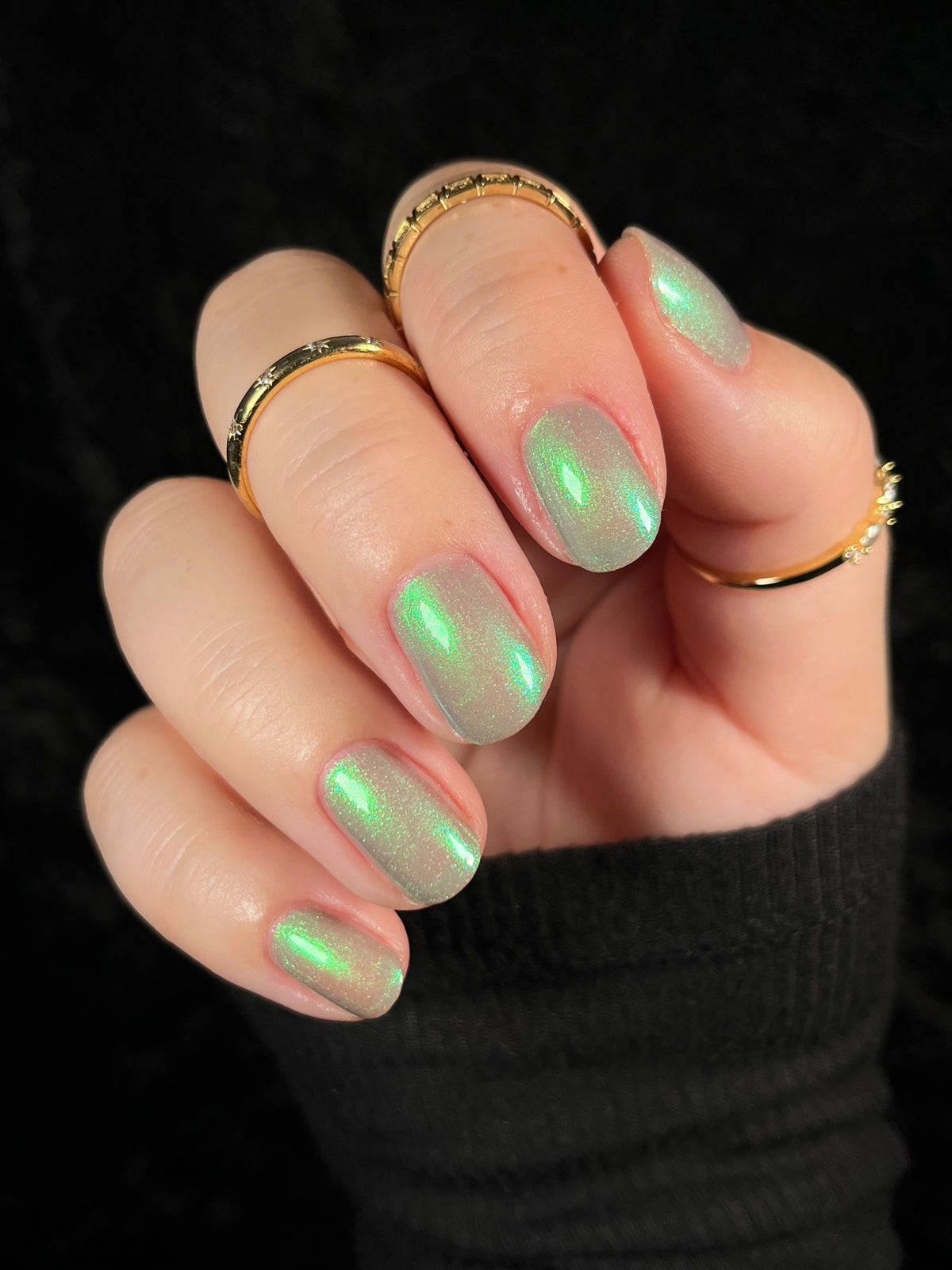 A close-up of a hand with manicured nails painted in a shimmering light green holographic polish, wearing several gold rings against a black background.