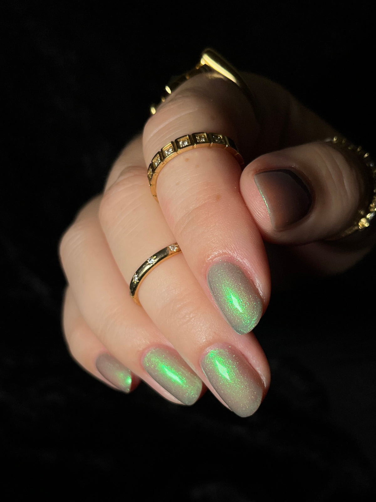 A close-up of a hand with manicured nails painted in a shimmering iridescent green polish, wearing several gold rings against a black background.