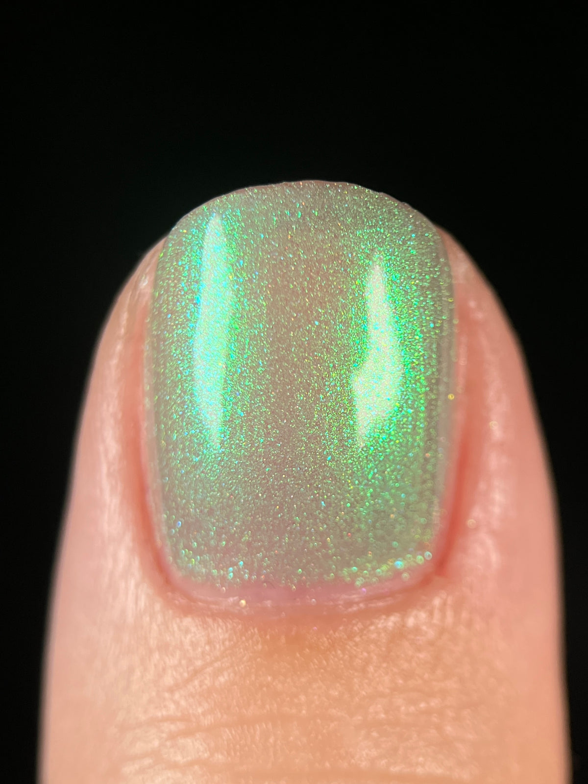 A close-up of a fingernail painted with a shimmering light green nail polish featuring fine glitter and a subtle iridescent sheen, set against a solid black background.
